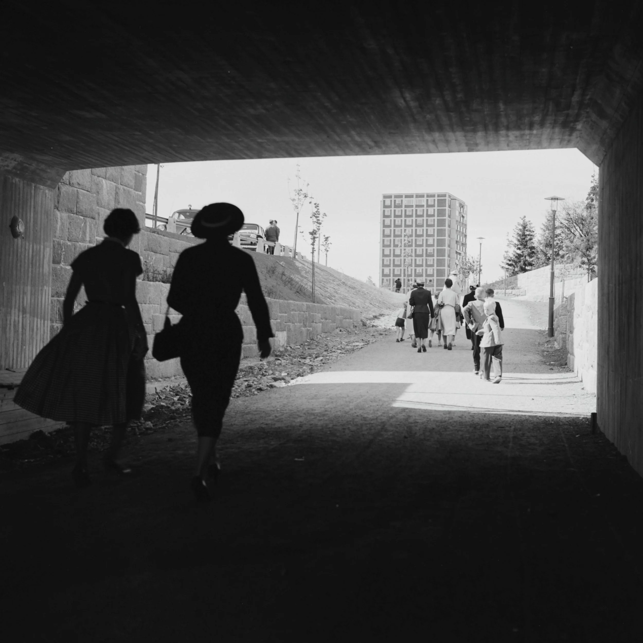 Siluett av två kvinnor under en gångtunnel, framför dem en grupp människor på gångvägen och ett höghus.