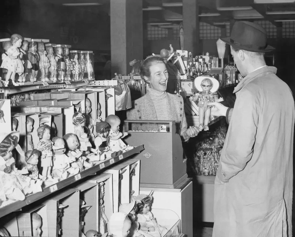 En kvinna vid en kassa och en butikshylla med dockor ler mot en manlig kund som håller upp en docka