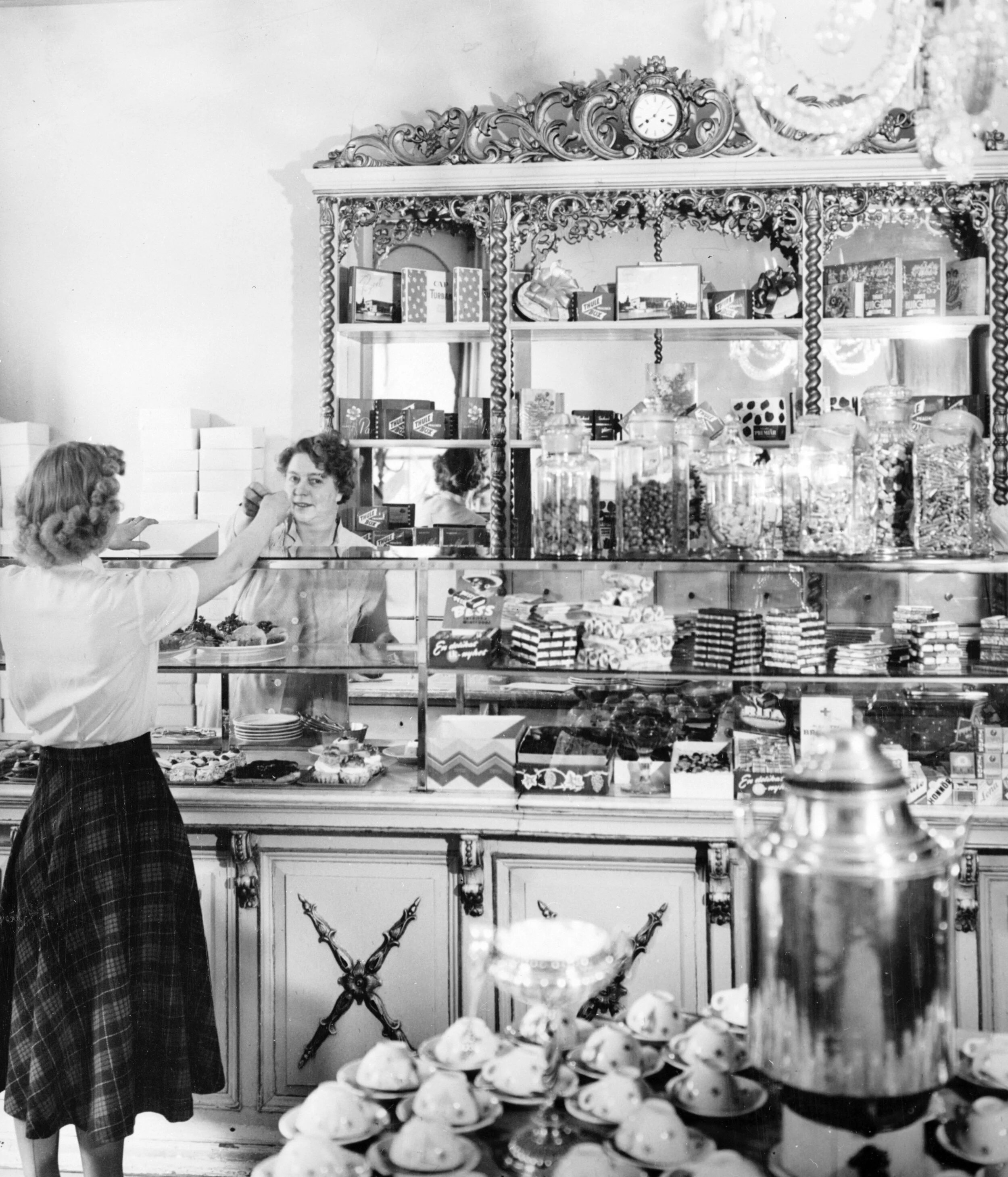Svartvitt foto inifrån ett konditori. På disken står fyllda glasburkar och kakor. I förgrunden syns en kaffetermos och uppochnervända koppar på fat. En person i rutig kjol betalar, personalen bakom disken räcker över växel och tittar in i kameran.