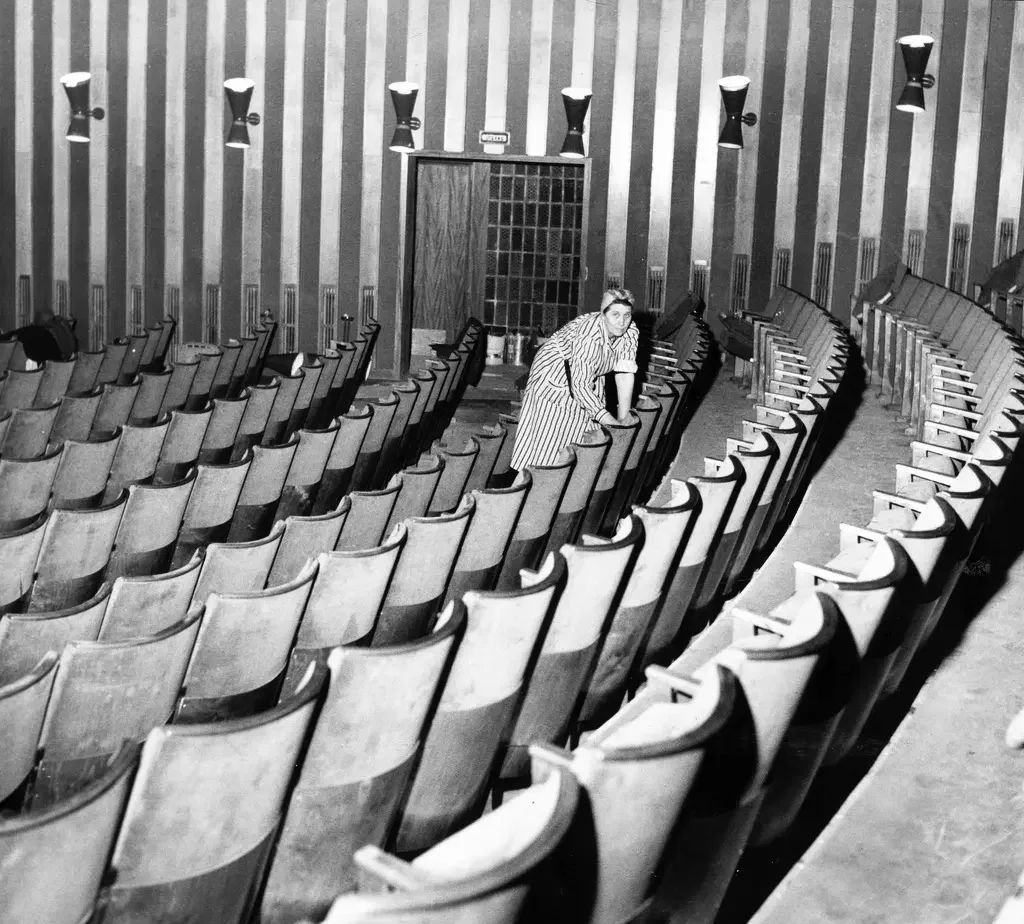 Kvinna står böjd över en stol i en biosalong iklädd randig rock och sjal på huvudet