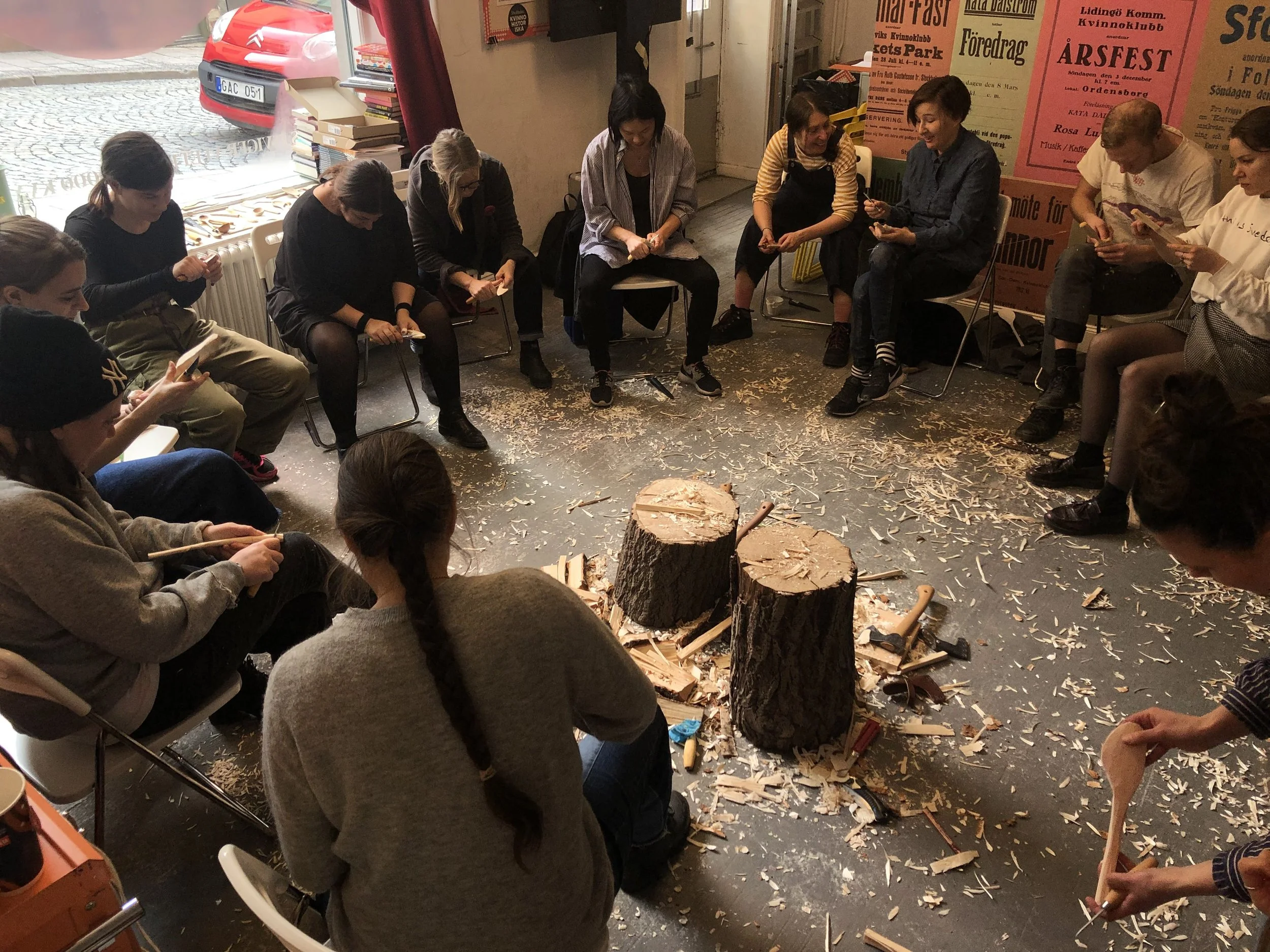 Workshop i täljning med två stubbar i mitten och en massa flisor på golvet, deltagarna sitter i en ring och täljer.