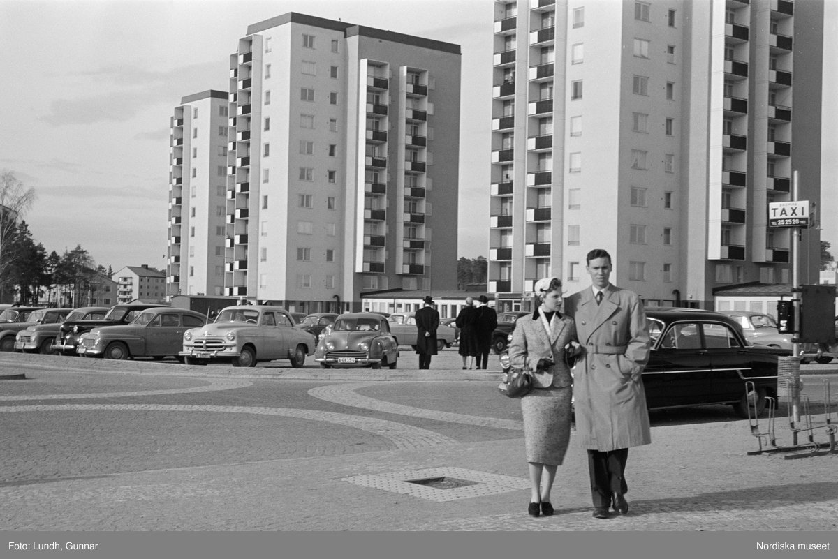 En man i rock och en kvinna i kjol och kavaj promenerar över ett torg arm i arm