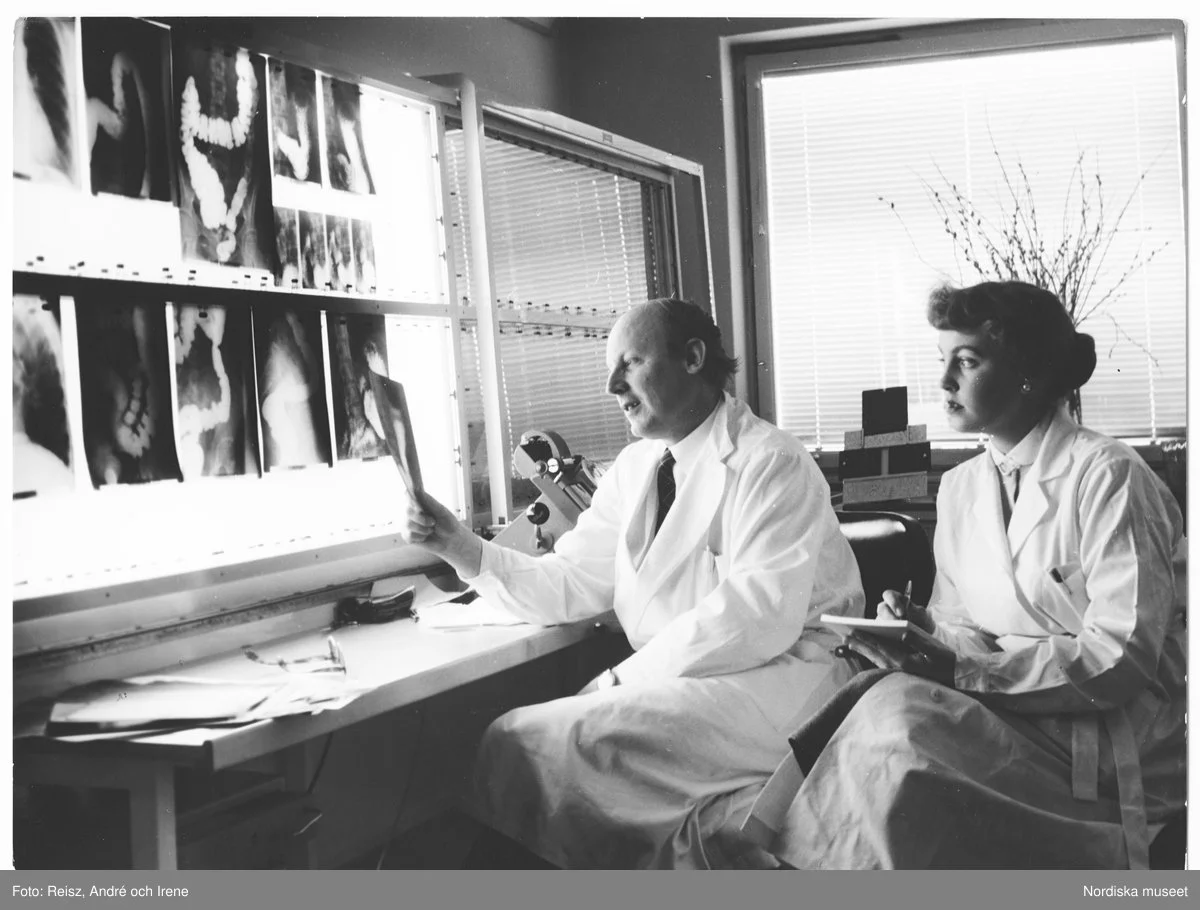 En manlig läkare i vit rock håller upp en röntgenbild, bredvid honom sitter en kvinna i vit rock med ett anteckningsblock och penna i handen och sneglar på bilden över hans axel