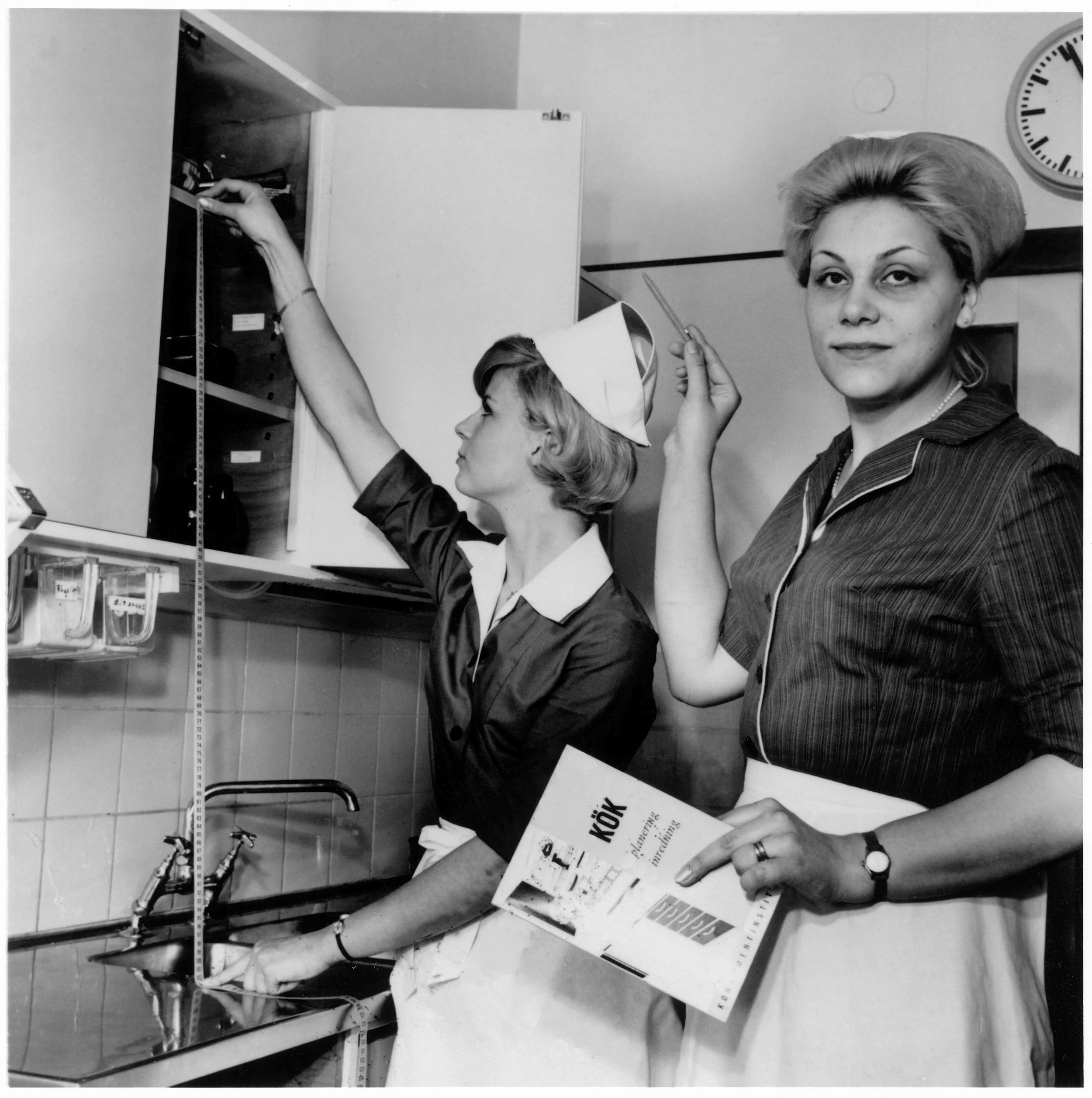 Två kvinnor i förkläde, blus och hatt står vid ett kök, den ena håller upp ett måttband och mäter avståndet mellan bänk och översta hyllan, den andra tittar in i kameran och håller i en penna och en broschyr.