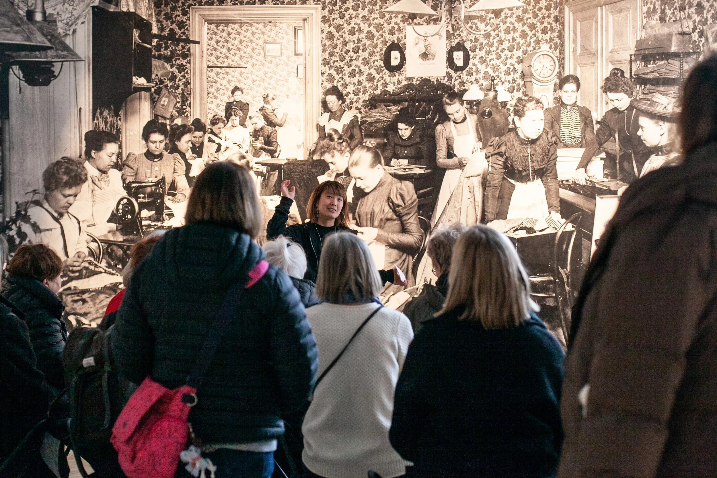 En grupp personer sitter framför en fondvägg av en arkivbild, där en person står och pekar och berättar