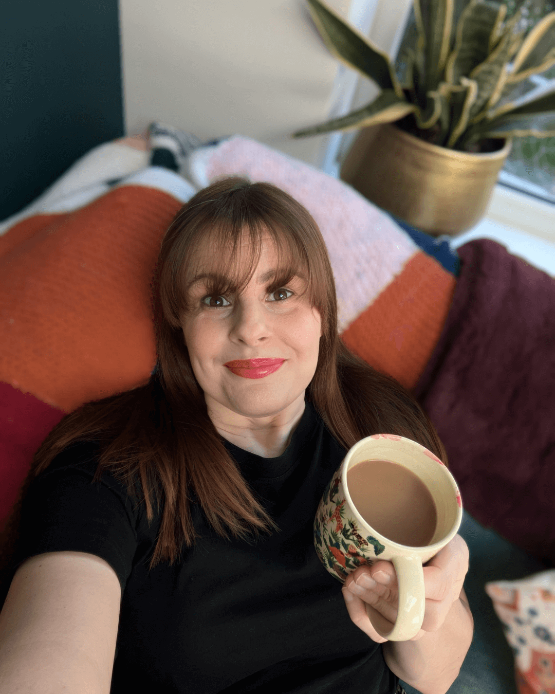 white woman with a fringe wearing a black top and red lippy sits with holding her cup of tea. There are blankets and a potted plant behind her.