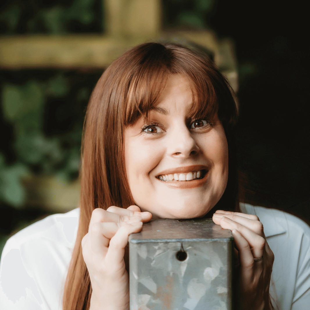 Kath grabs onto a metal fence post and rests her chin on the top. She's smiling and looking straight into the camera.