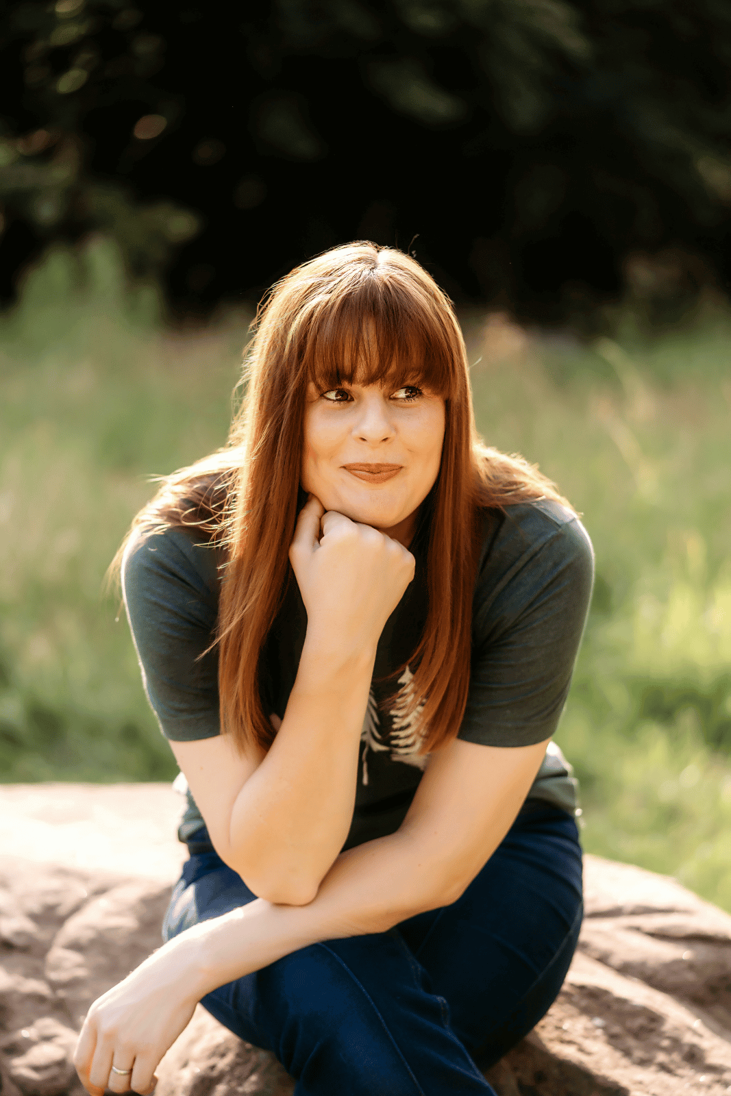 Kath is sitting on a massive rock in the woods. She's resting her chins on her hand and looking up to the right of the camera. She's wearing a green t-shirt with white trees on it.