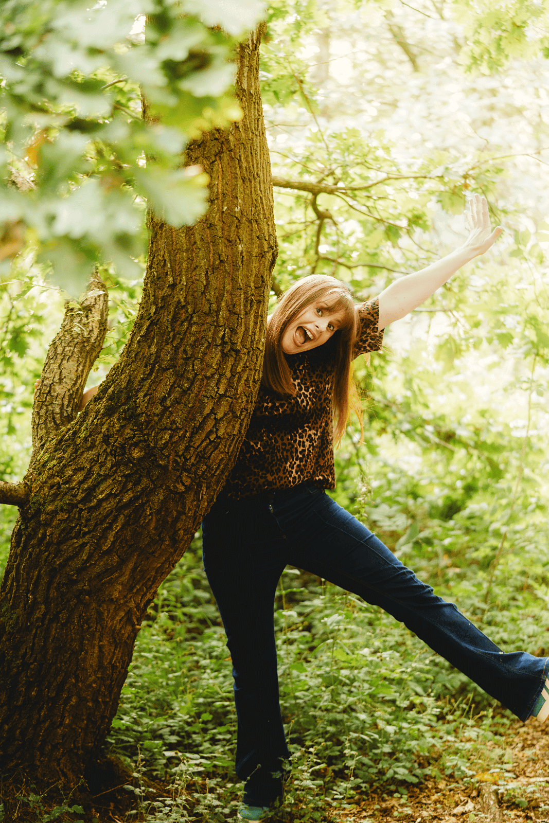 white woman leaning on a tree with her arm and leg out in a star shape and a goofy grin on her face. She's wearing jeans and a leopard print tshirt