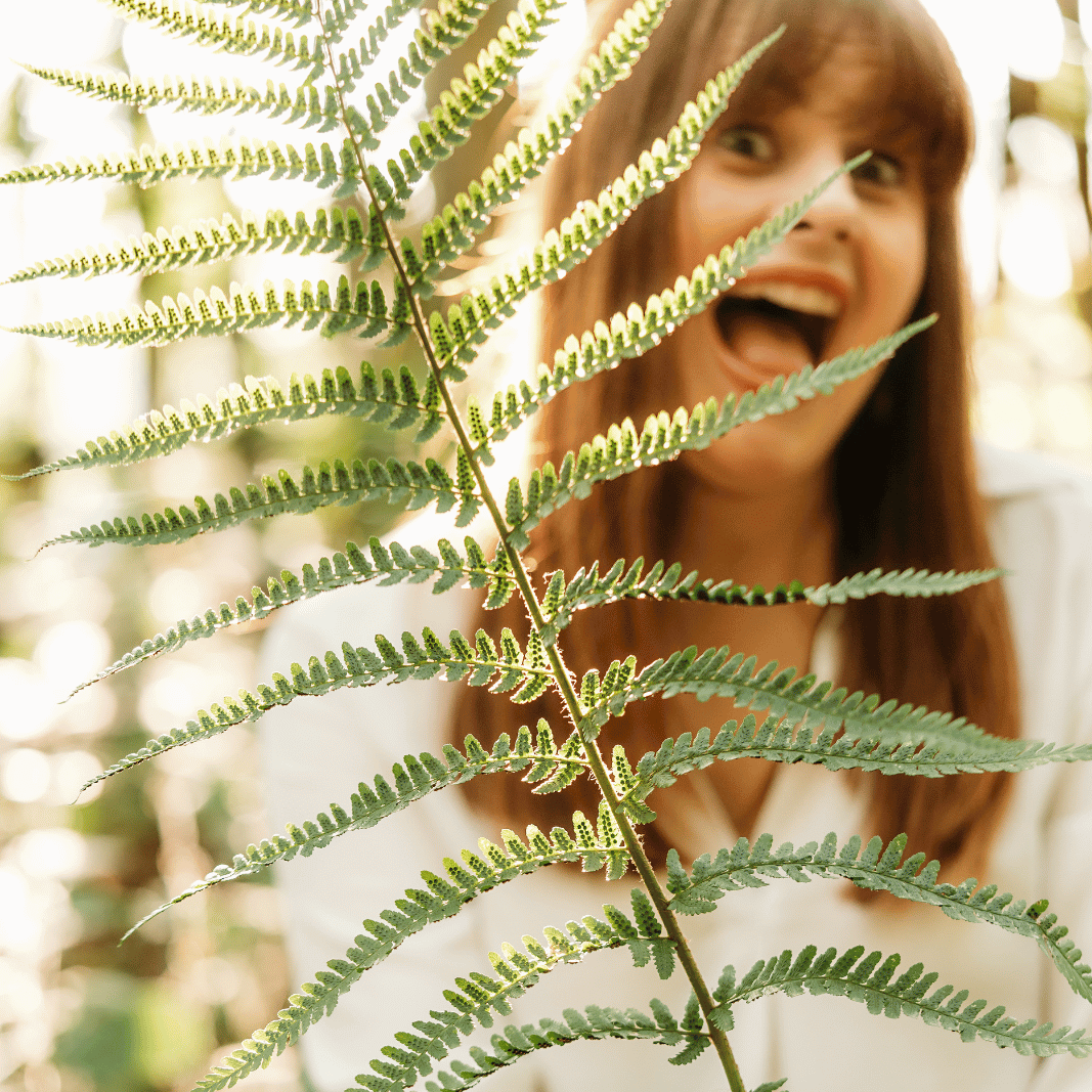 white woman wearing a white shirt holds up a leaf. The leaf is in focus and her face is blurred out, but you can still tell she's making a happy, goofy face