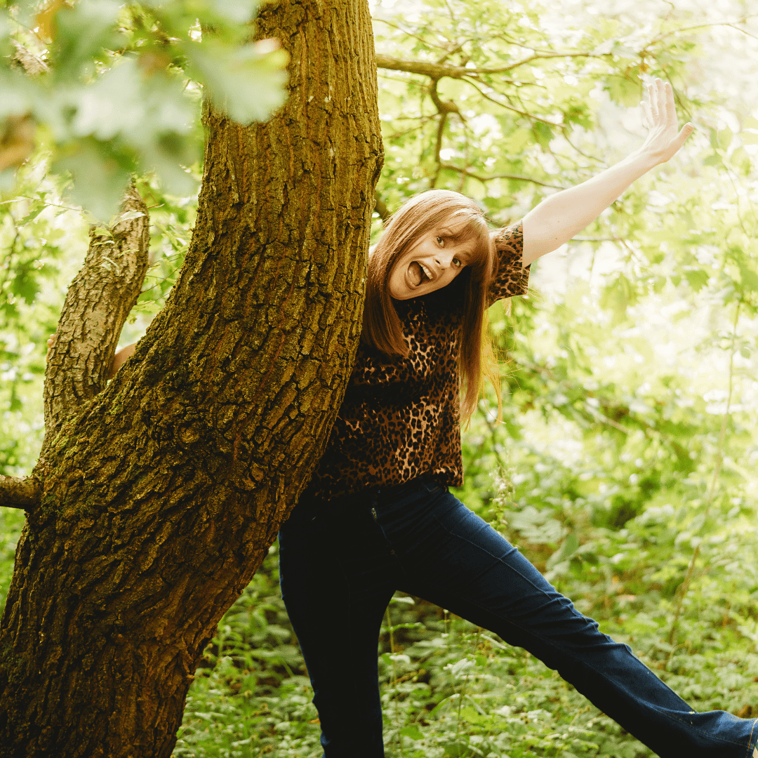 Kath is leaning on a tree while making a star shape with her arm and leg. We don't know why, but she looks happy. She's wearing jeans and a leopard print tee.