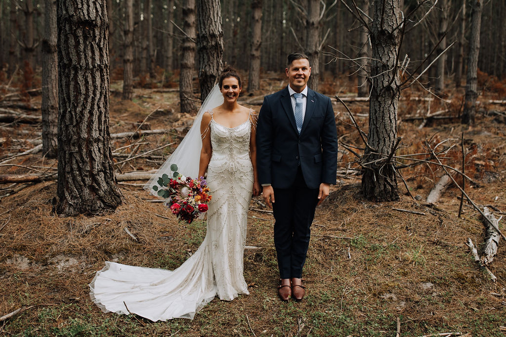 Carla and rob holding a bouquet of cute flowers with a white gown in the woods