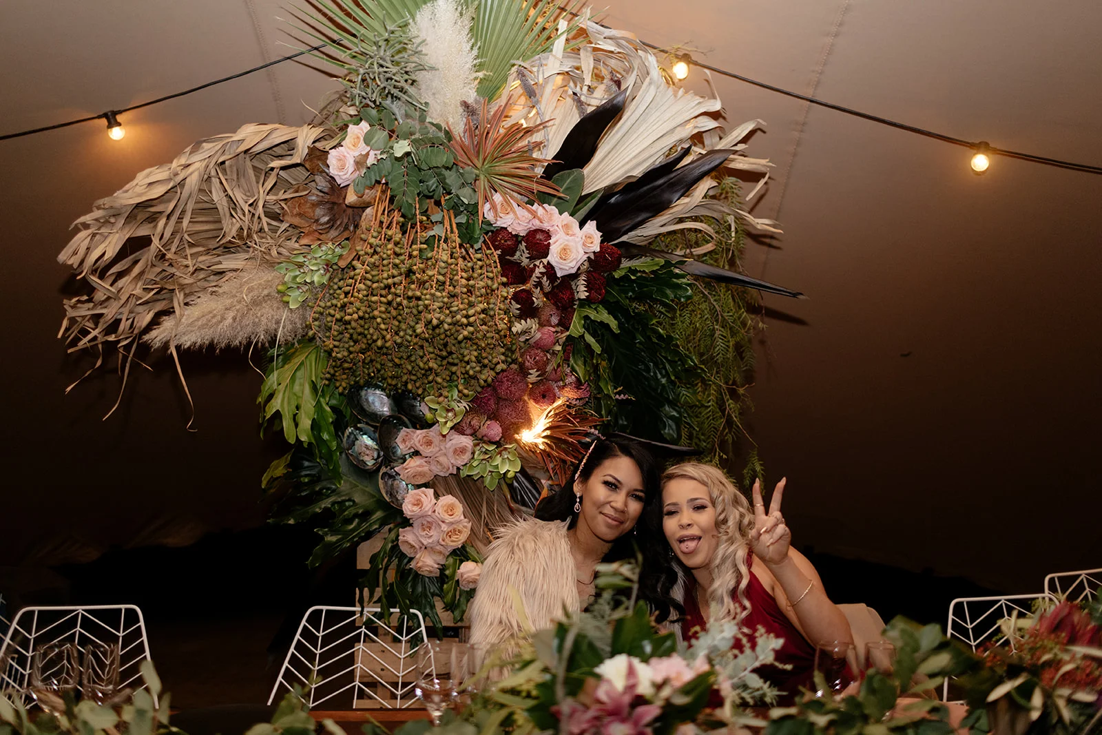 bride at the wedding reception seated on a table full of flowers posing with friend
