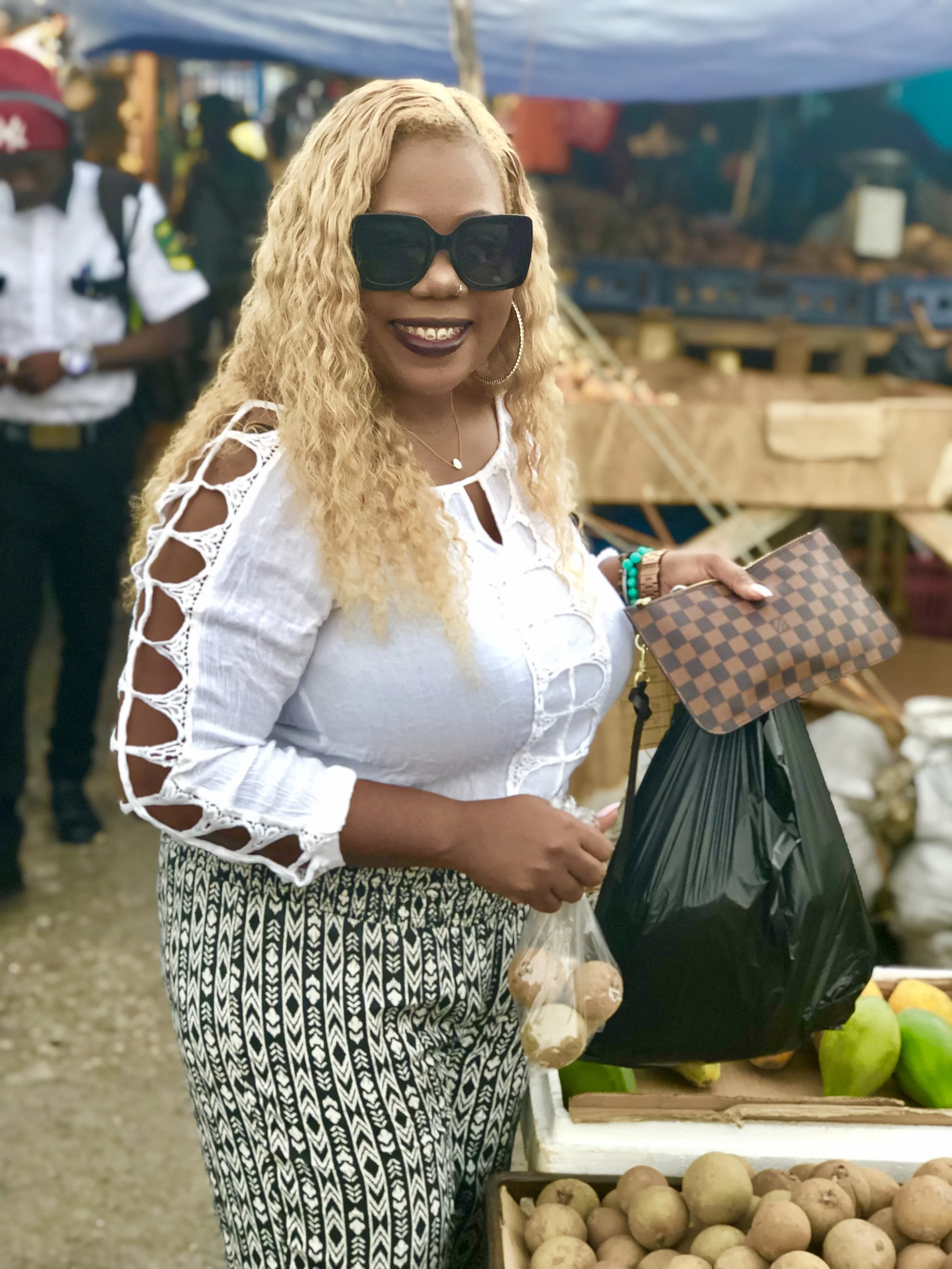 Woman with blonde curly hair, wearing large dark sunglasses, a white top with cutout sleeve details, and patterned pants, holding a checkered clutch and a black shopping bag at a market stall with produce.