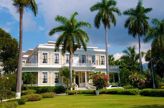 A large, multi-story house with white exterior and numerous windows, surrounded by lush green lawn and tall palm trees under a partly cloudy sky.