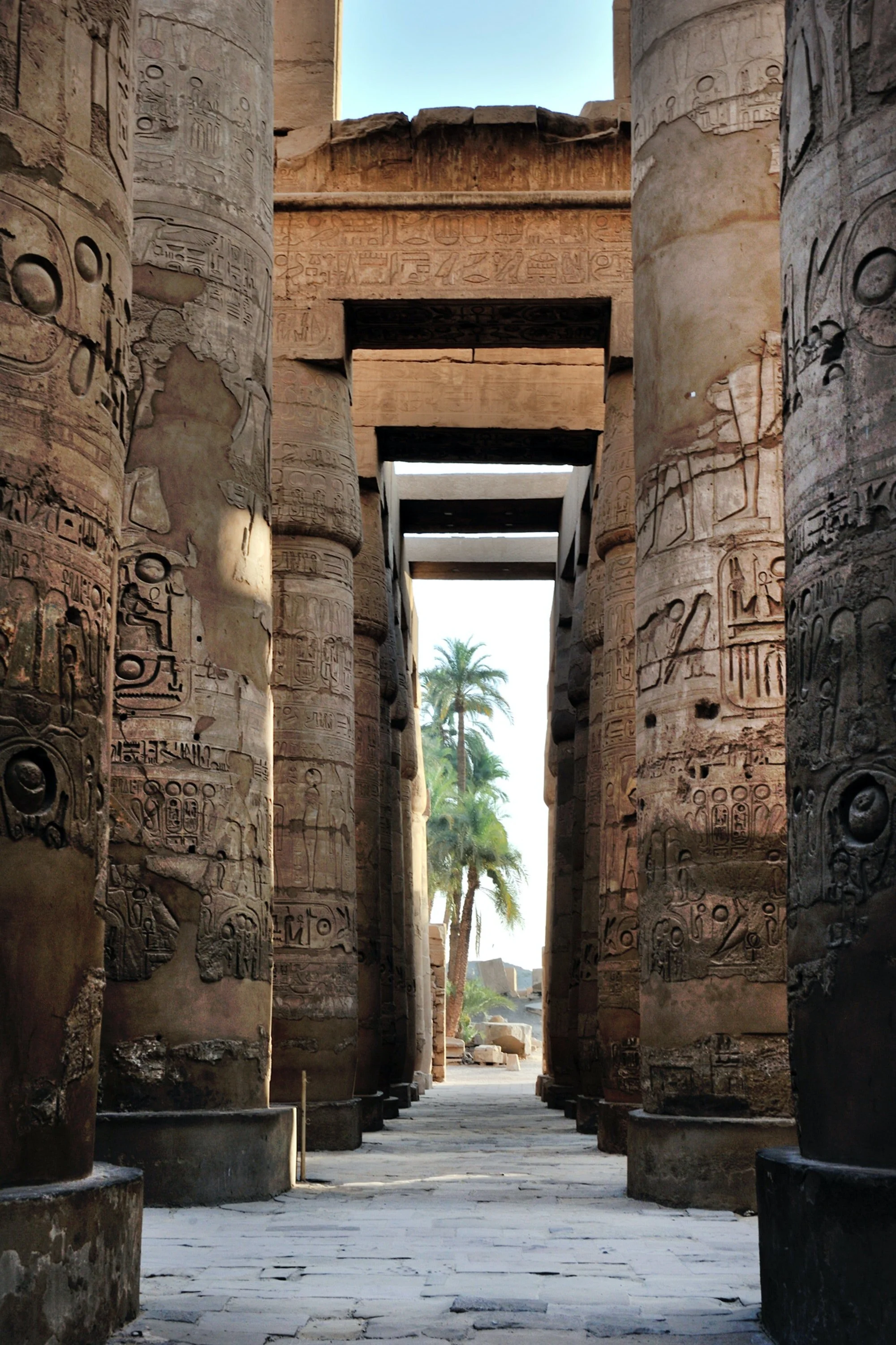 Ancient Egyptian stone pillars with hieroglyphics inscribed, leading to an open area with palm trees visible in the background.