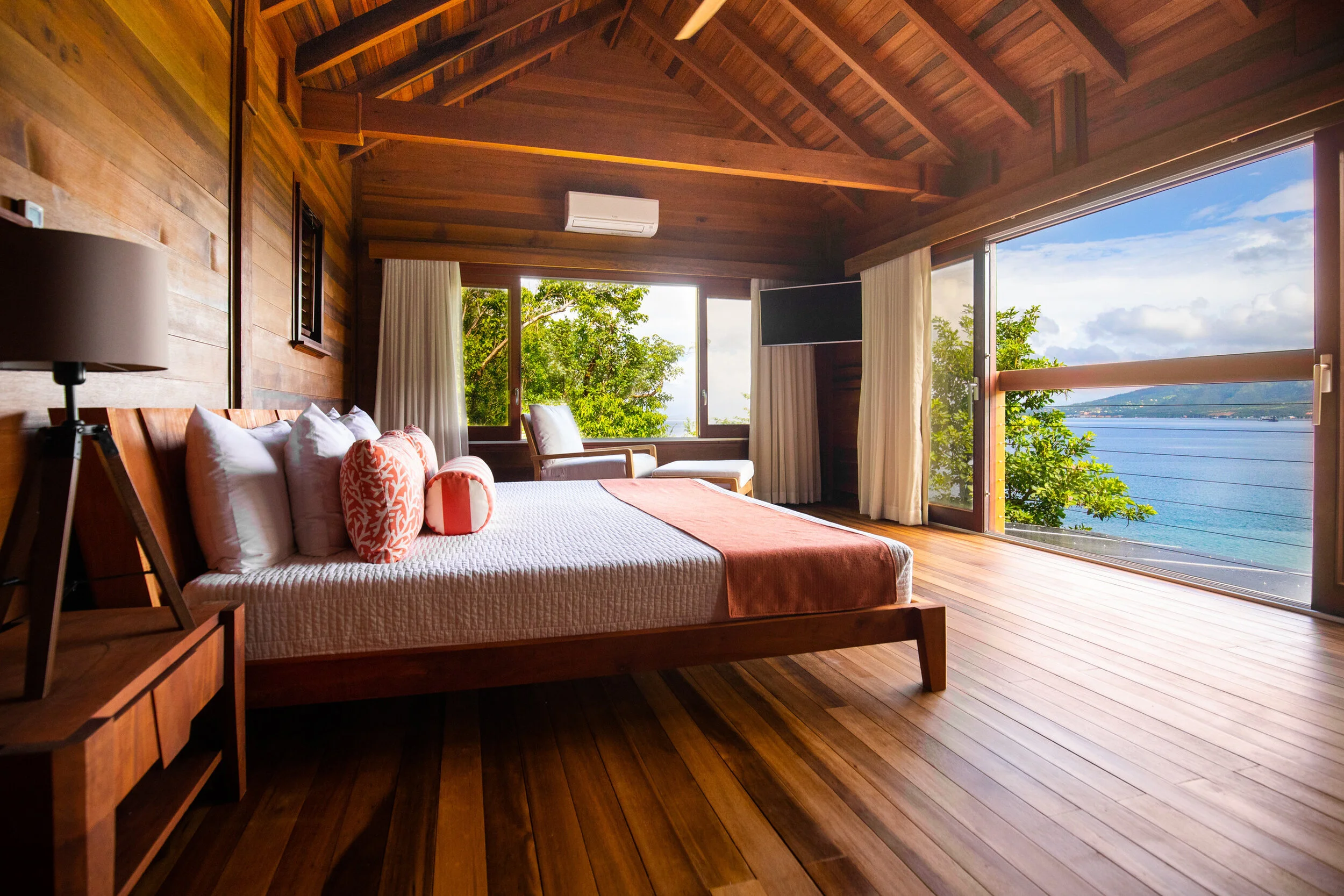 Bedroom with wooden walls and ceiling, large windows showing greenery and water, bed with white bedding and orange accents, and a chair facing outward.