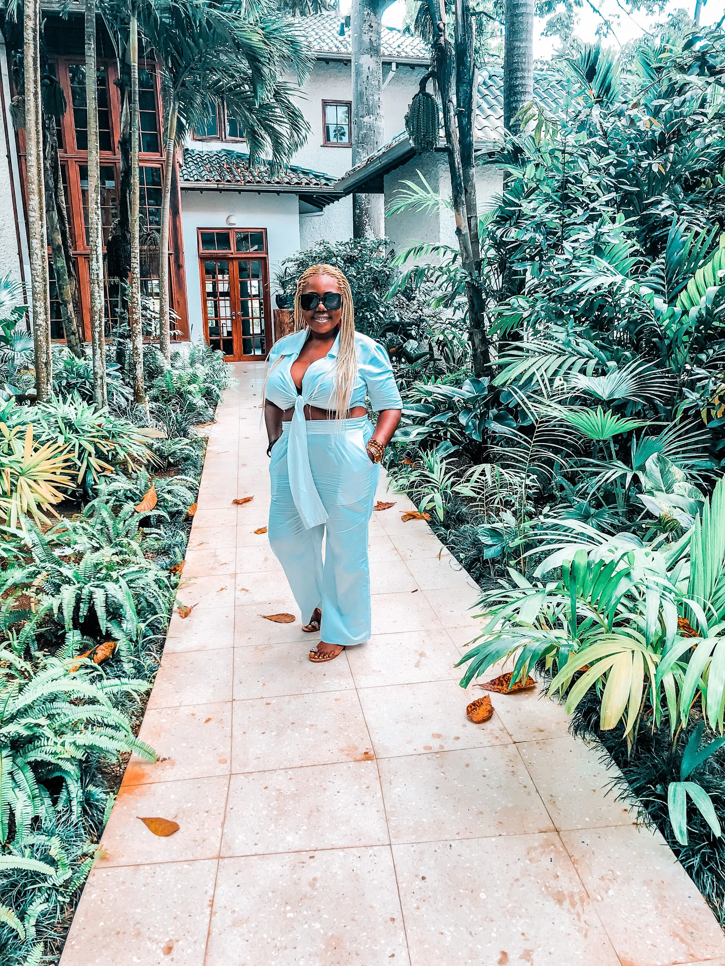 A woman with blonde braids, wearing black sunglasses, a light blue outfit, with a tie-front top and wide-leg pants, stands on a tiled garden walkway surrounded by lush tropical plants and trees, with a white house with wooden windows and doors in the background.