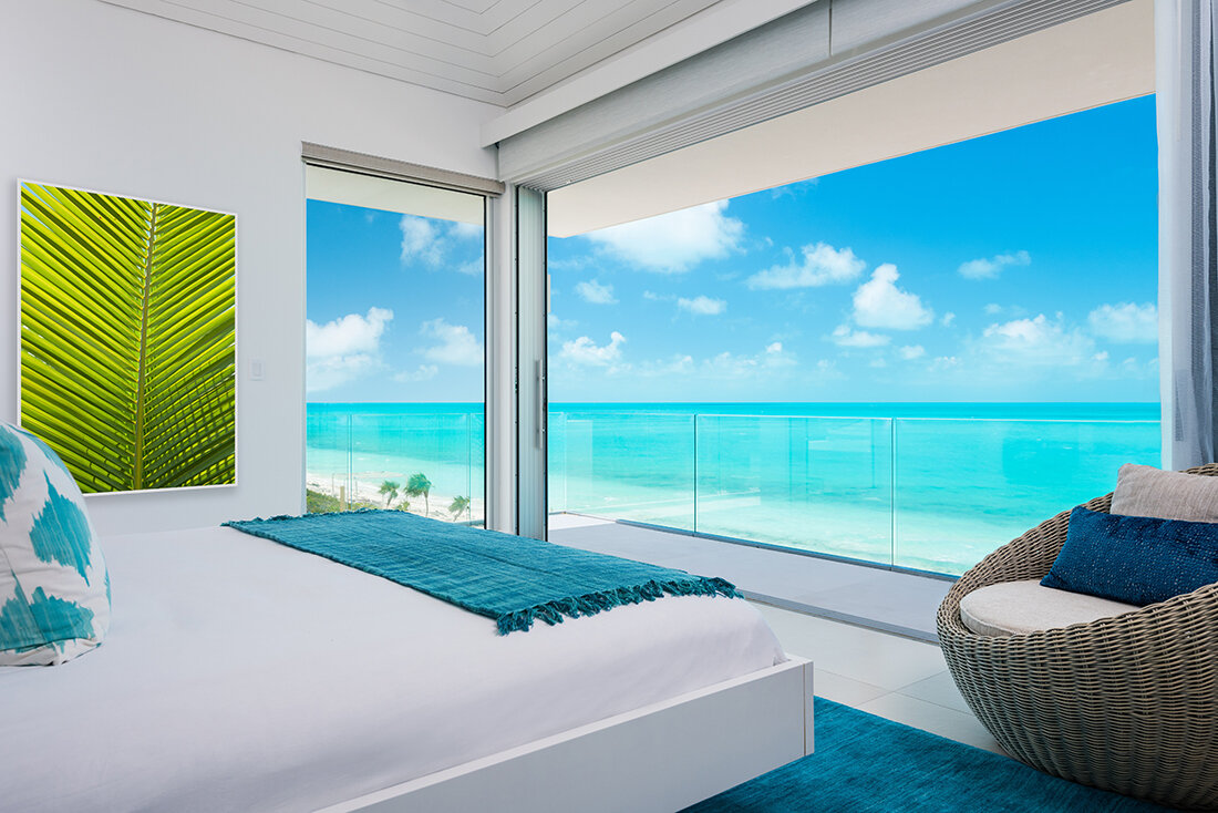 A modern bedroom with a large window and balcony overlooking a beach and the ocean. The room has a white bed with blue and white pillows, a blue throw, a wicker chair, and a green leaf art piece.
