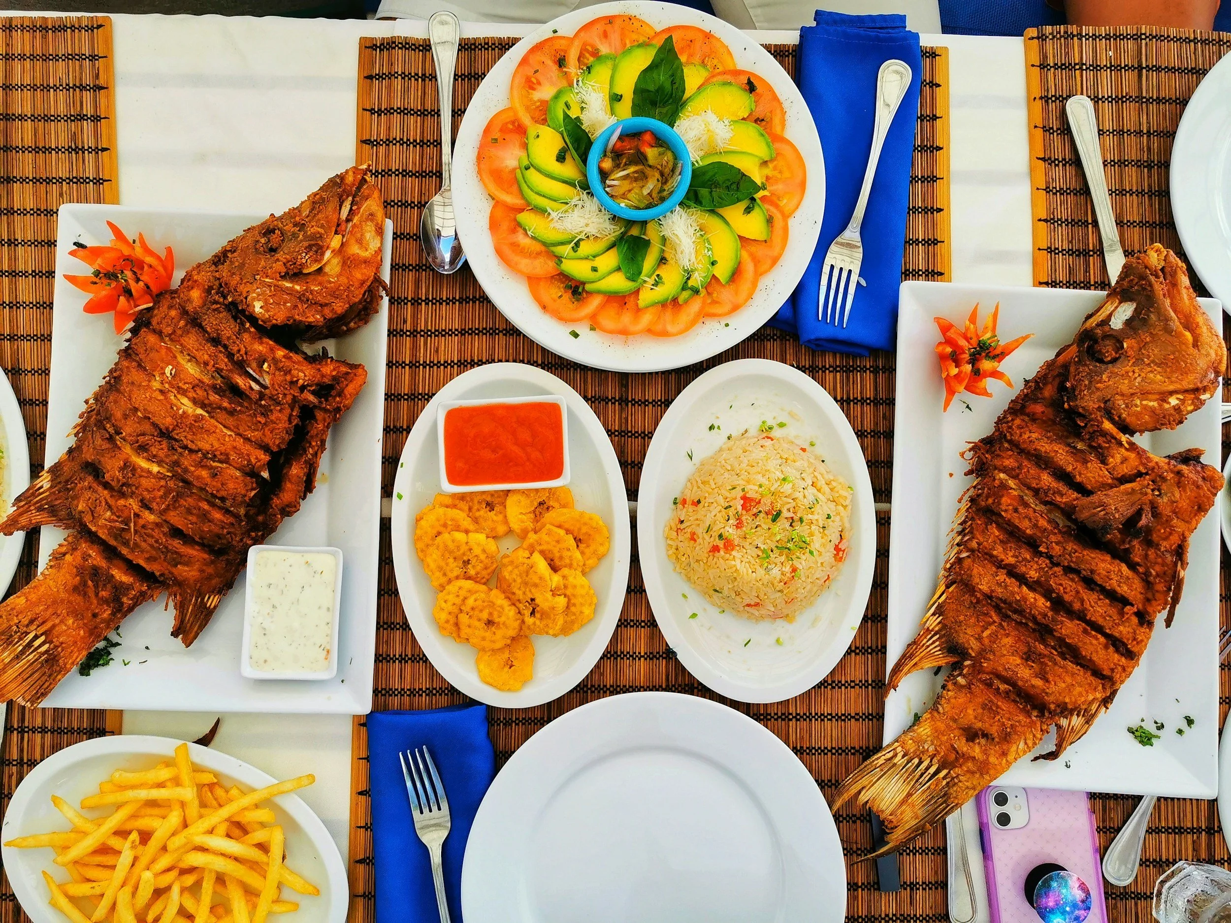 A top-down view of a table with various dishes including whole fried fish, a salad with tomato, avocado, and basil, French fries, rice, and dipping sauces.