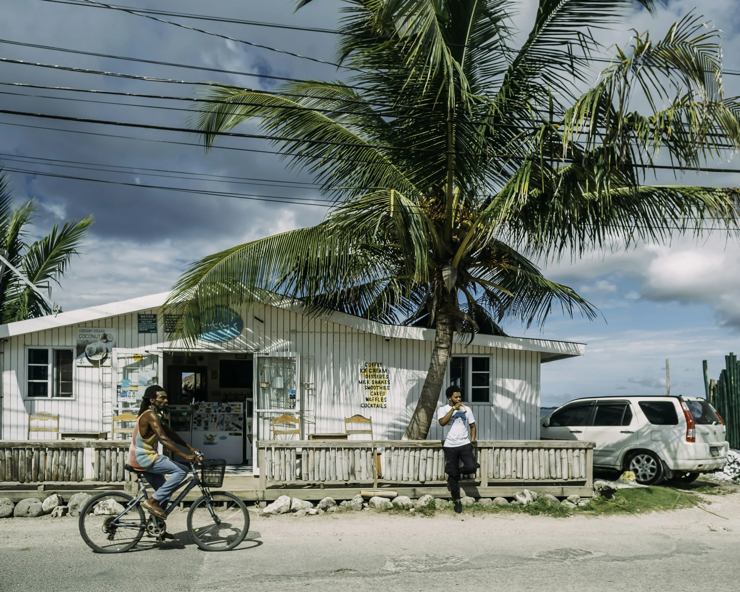 A white building with a small open window and a sign listing menu items such as coffee, ice cream, desserts, milkshakes, smoothies, cakes, waffles, and cocktails. In front, a man riding a bike, a woman walking, and a person with dark hair. A large pa