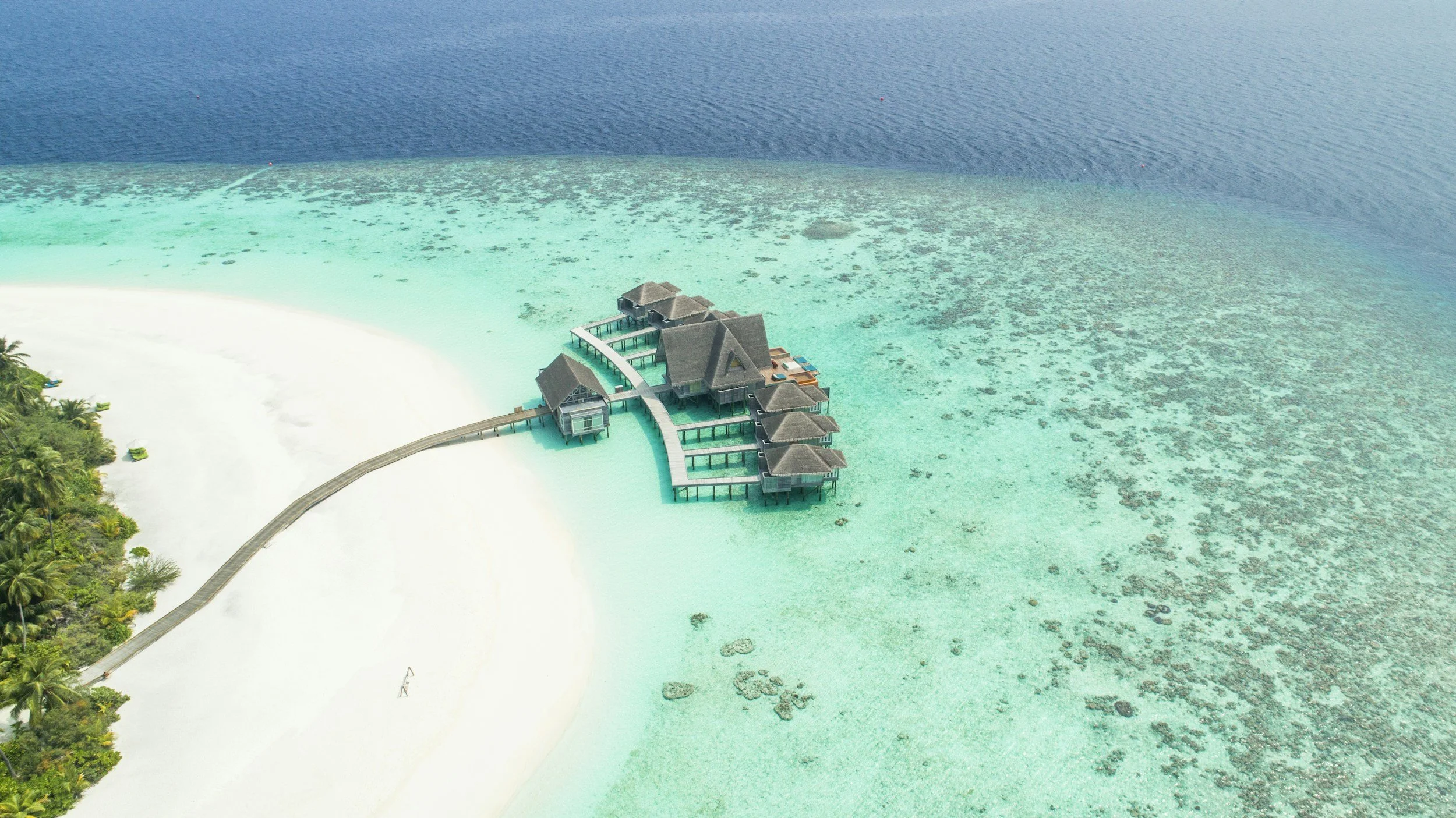 Aerial view of overwater bungalows on a turquoise beach with clear shallow waters and a dense green jungle on the left.