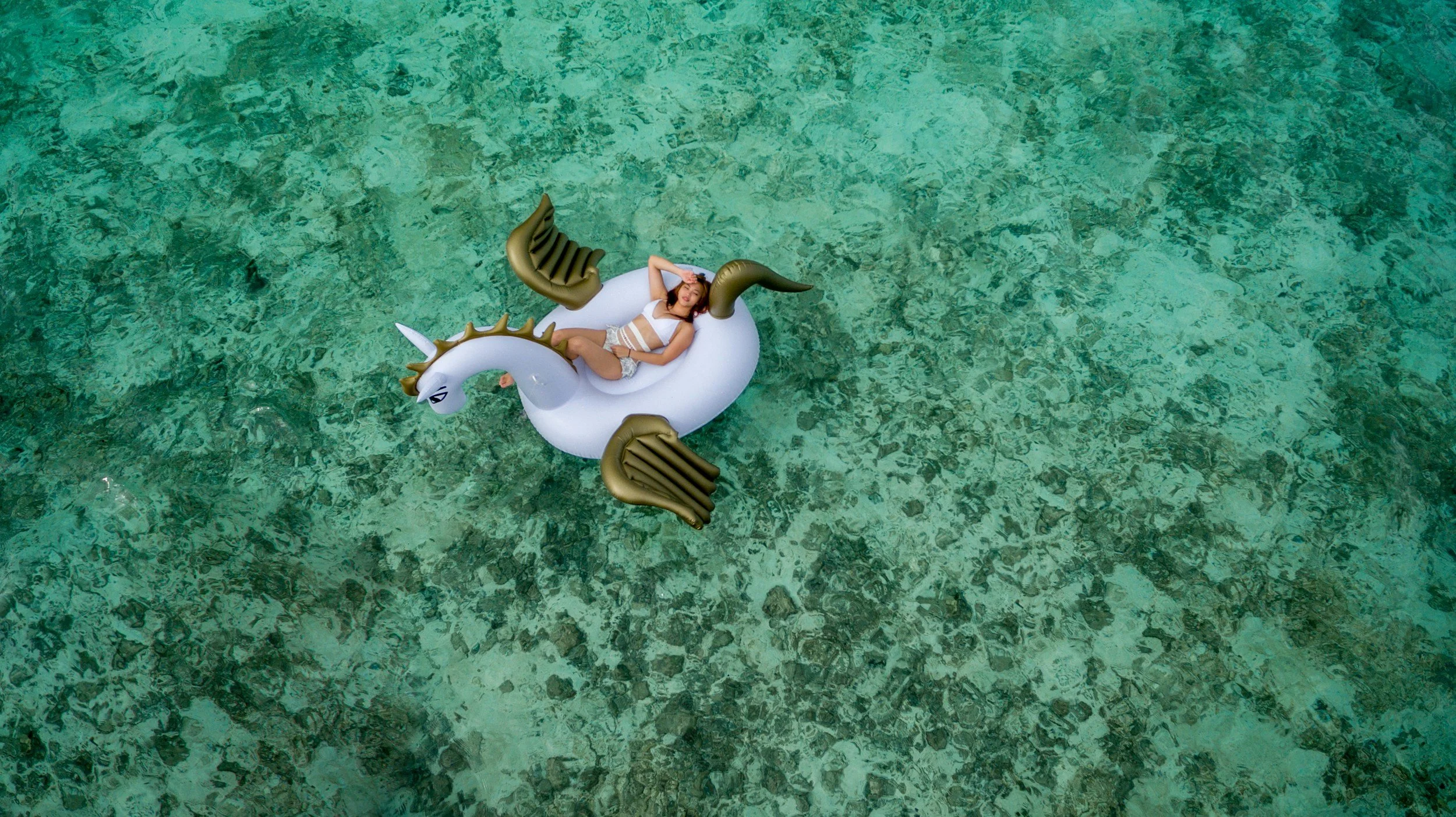 A woman relaxing on an inflatable unicorn float in clear, shallow water.