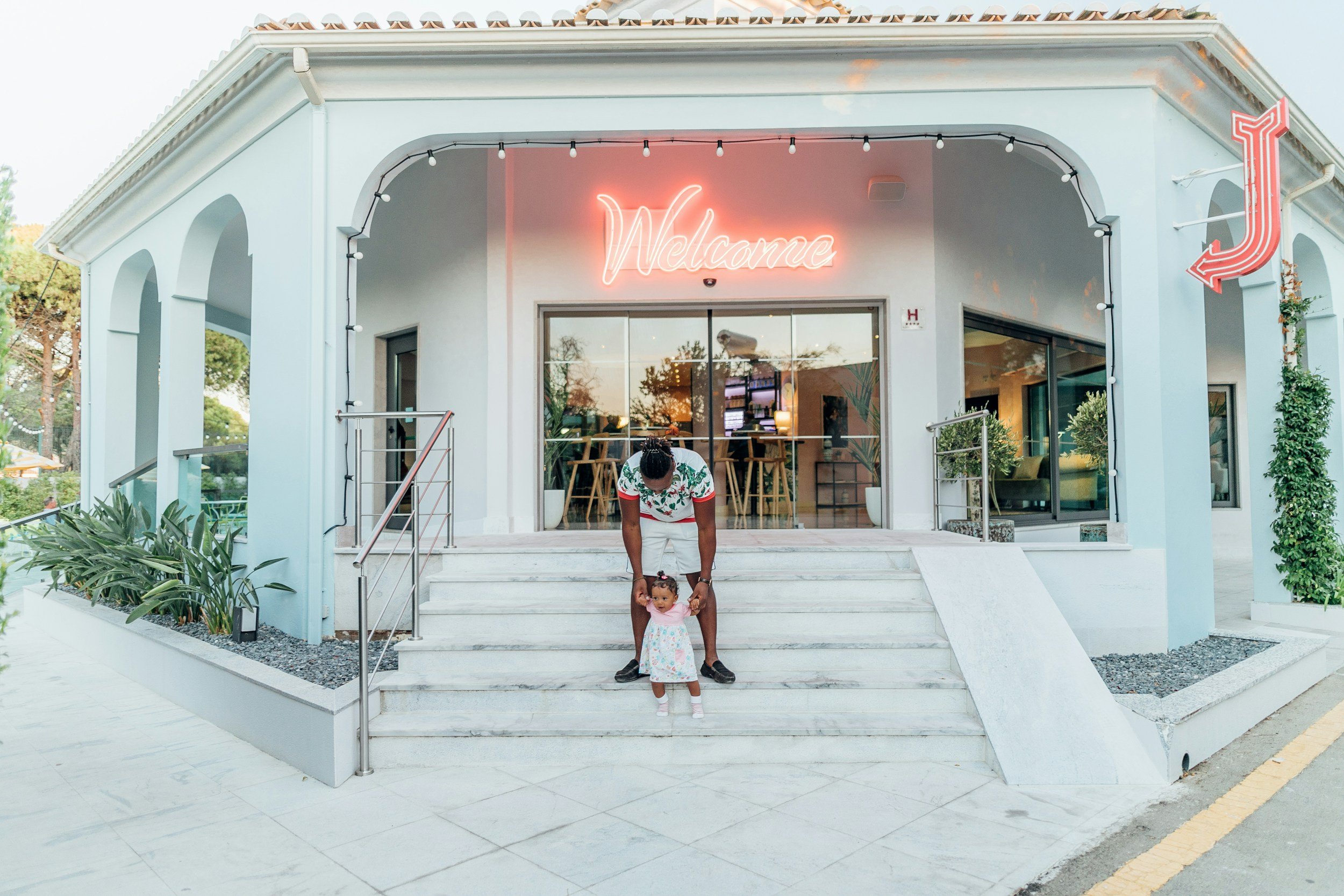 A man and a young girl are on the front steps of a light blue building with a neon 'Welcome' sign and a neon arrow. The man is holding the girl by her hands, and the girl is wearing a dress with a pink top and floral skirt. The building has large win
