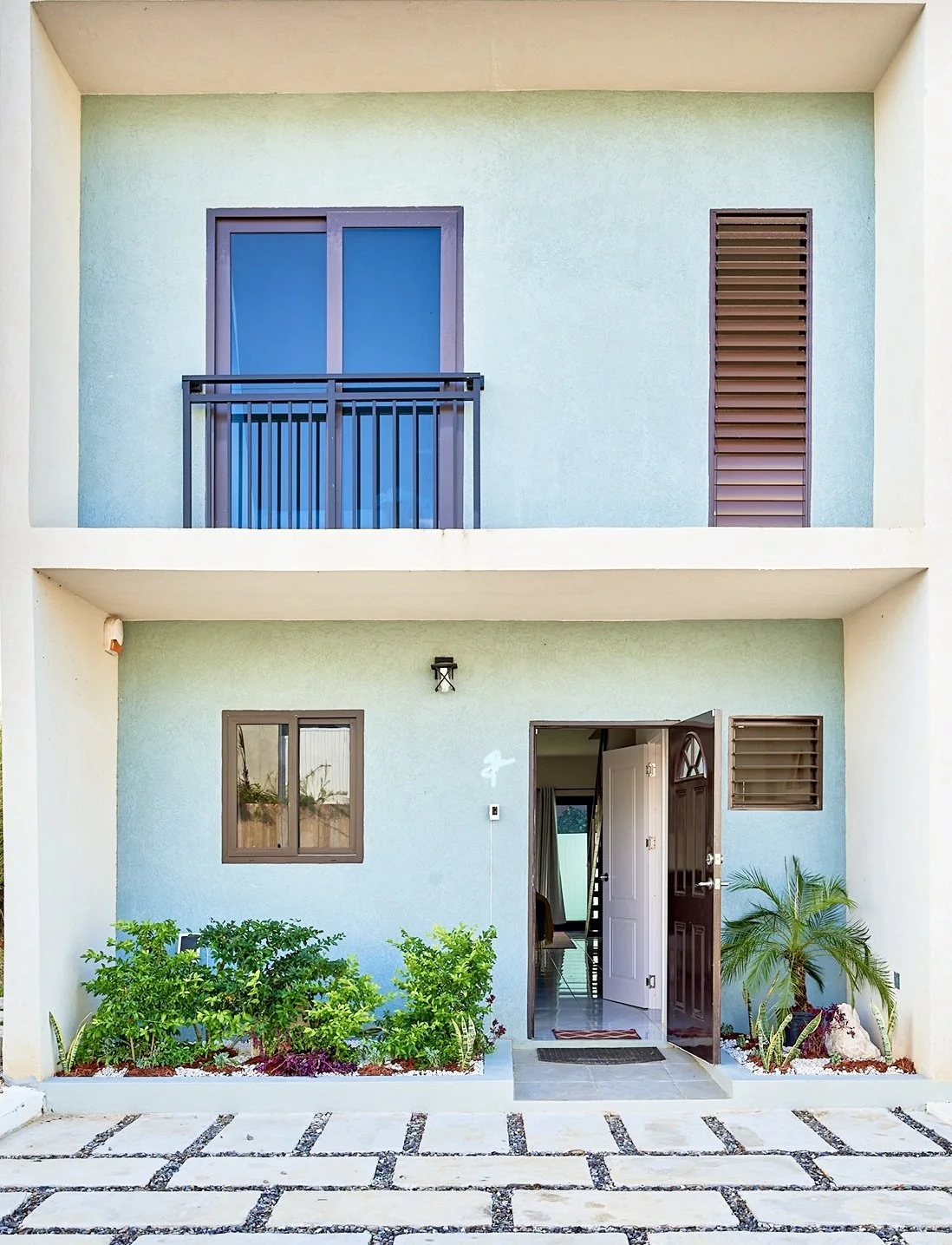 Front view of a modern two-story residential building with a light blue exterior. The ground floor features a door open to reveal the interior, flanked by two windows. The second floor has a sliding glass door with a small balcony and a window with b
