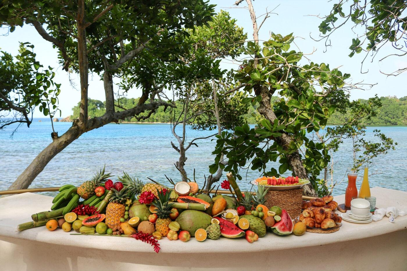 A table set with various dishes including a large plate of sliced tomatoes, avocado, basil, and shredded cheese, along with fried fish, french fries, rice, small bowls of dipping sauces, and condiments.