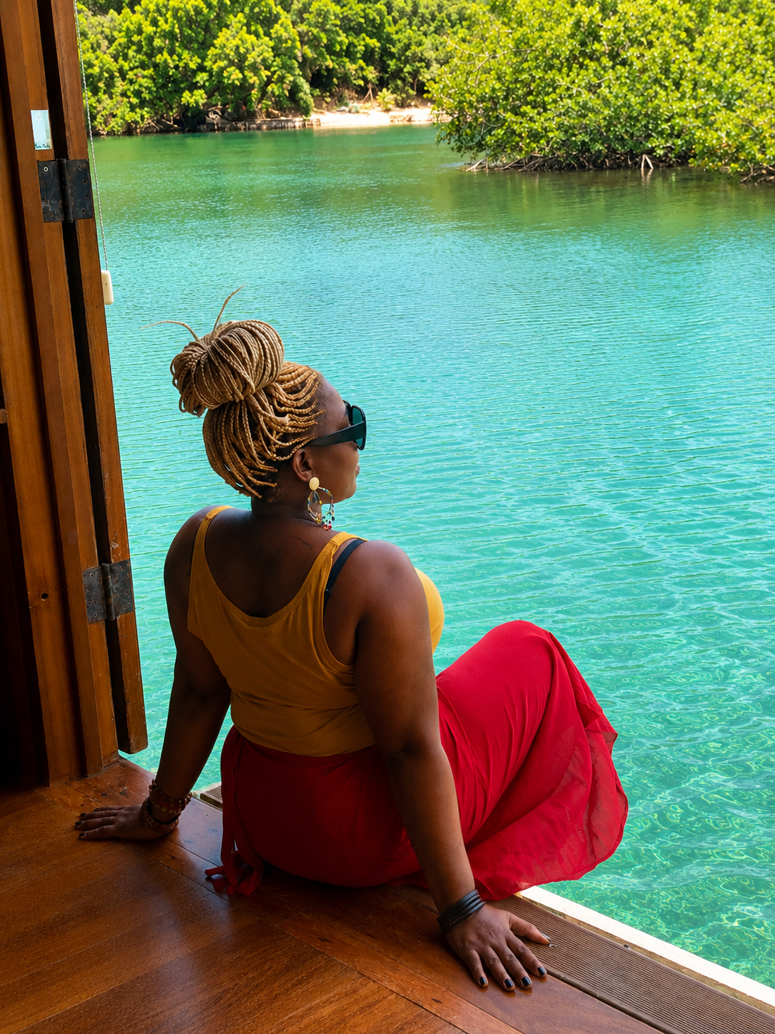 A woman with braided blonde hair, wearing sunglasses, a yellow tank top, and a red skirt, sitting at the edge of a wooden hut overlooking a turquoise river and green foliage.