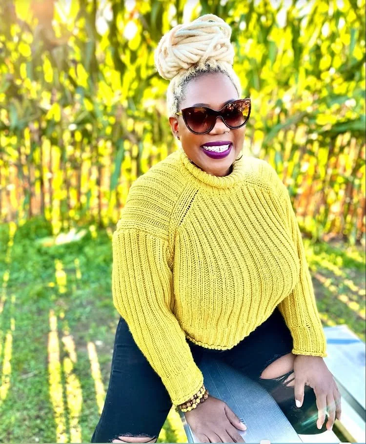 A woman with blonde dreadlocks in a bun, wearing large sunglasses, purple lipstick, a yellow knitted sweater, and black distressed jeans, sitting outdoors in a grassy area with tall yellow-green plants in the background.