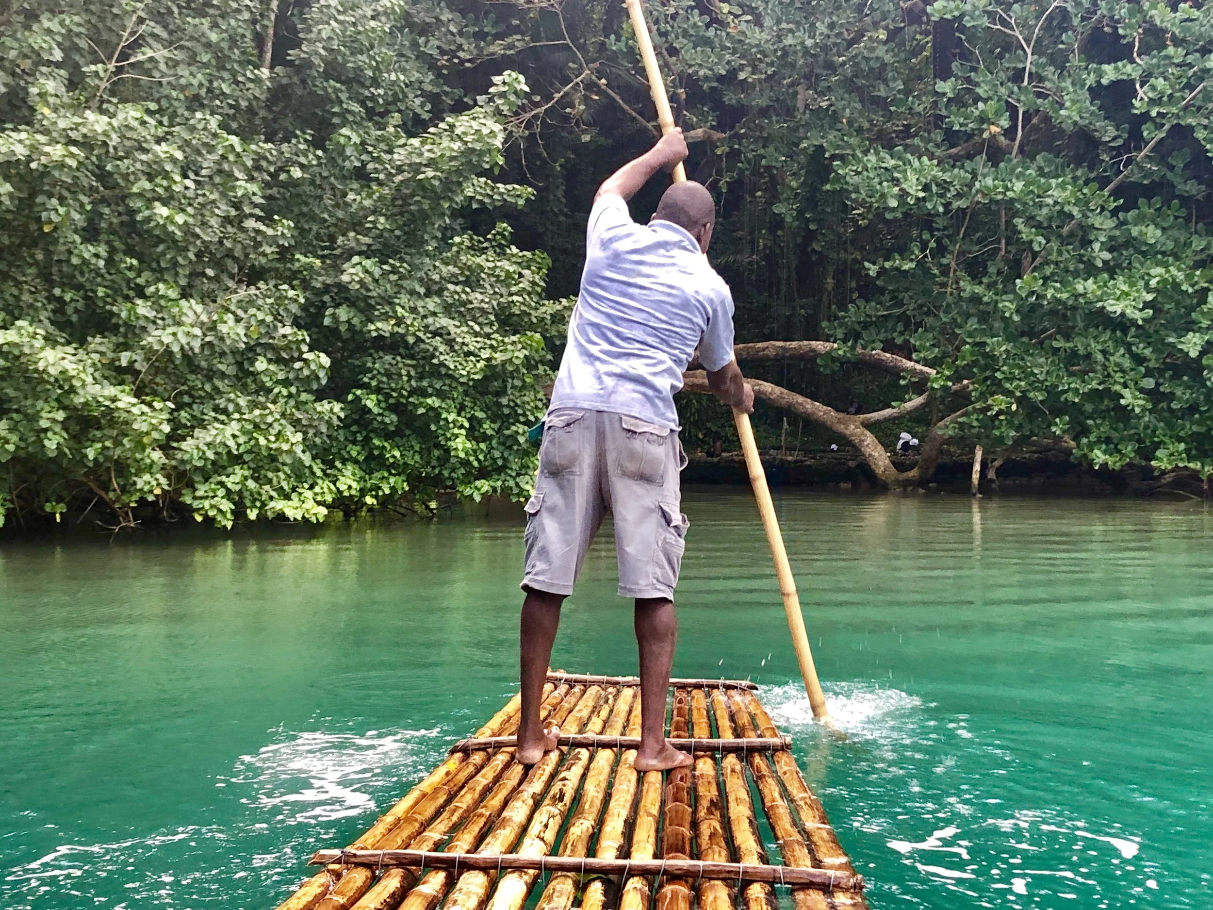 A person standing on a bamboo raft in a green river, surrounded by lush vegetation. The person is holding a long pole, using it to navigate the water.