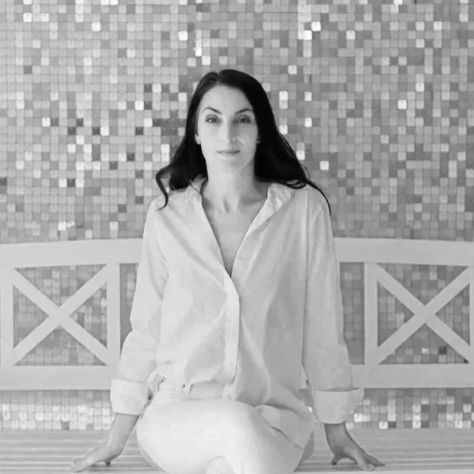A woman with dark hair wearing a white shirt sitting on a bench with a mosaic tile wall in the background.