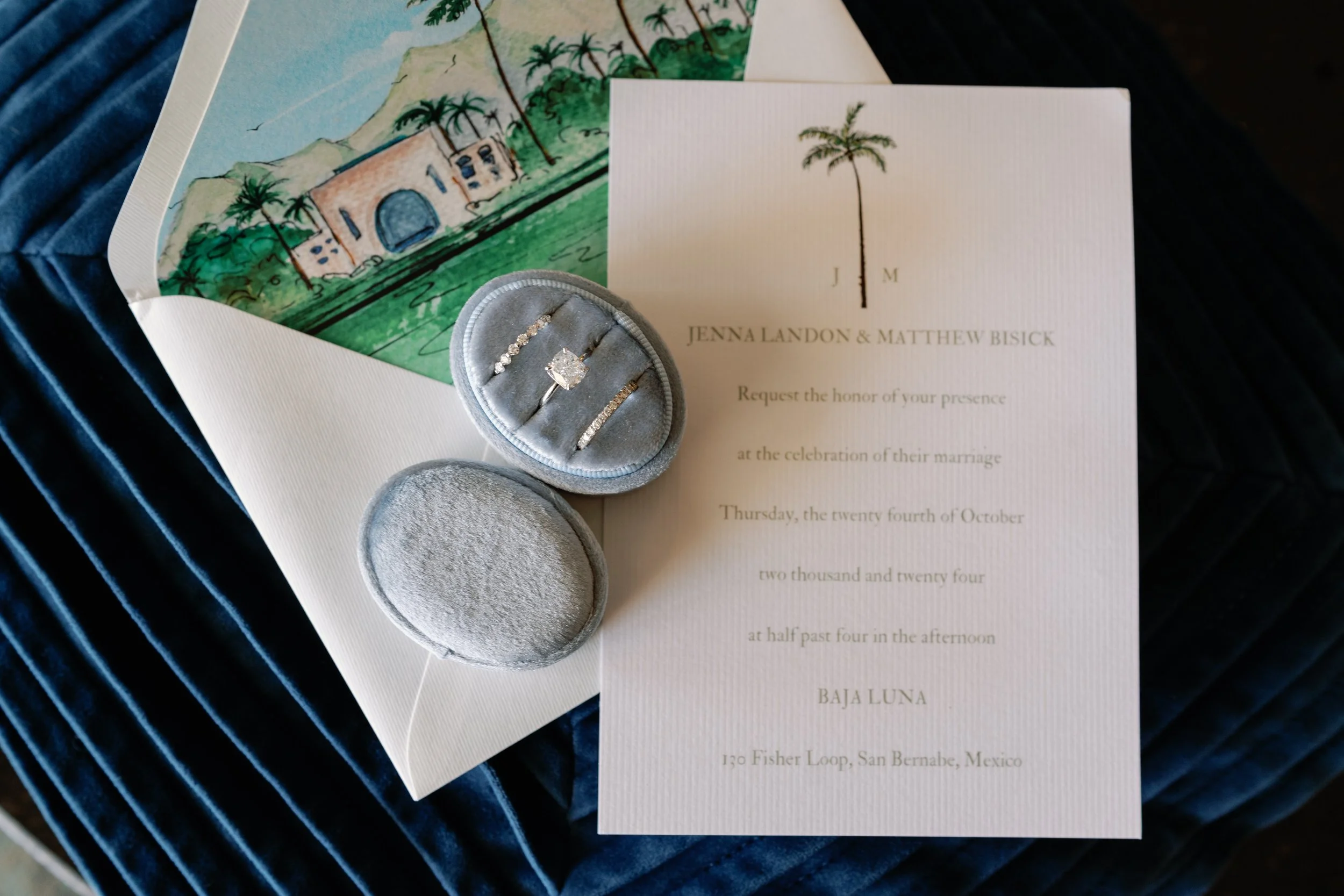 Wedding invitation with rings and jewelry box on dark blue velvet fabric. The invitation has a palm tree and Montezuma castle illustration, and the jewelry box contains a diamond engagement ring and wedding bands.