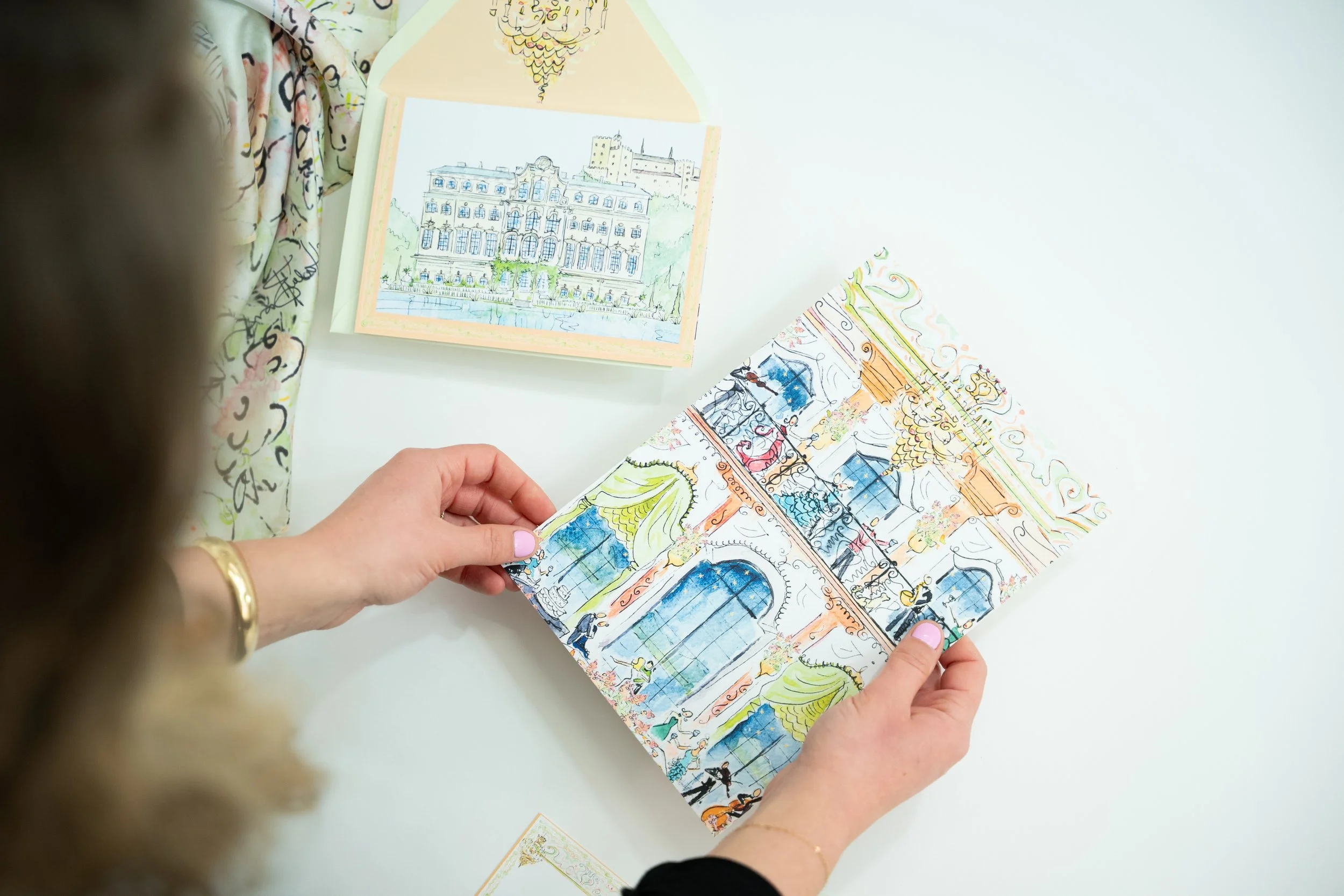 Person holding a colorful illustrated booklet or foldout with architectural sketches and drawings, with a hand, on a white surface.