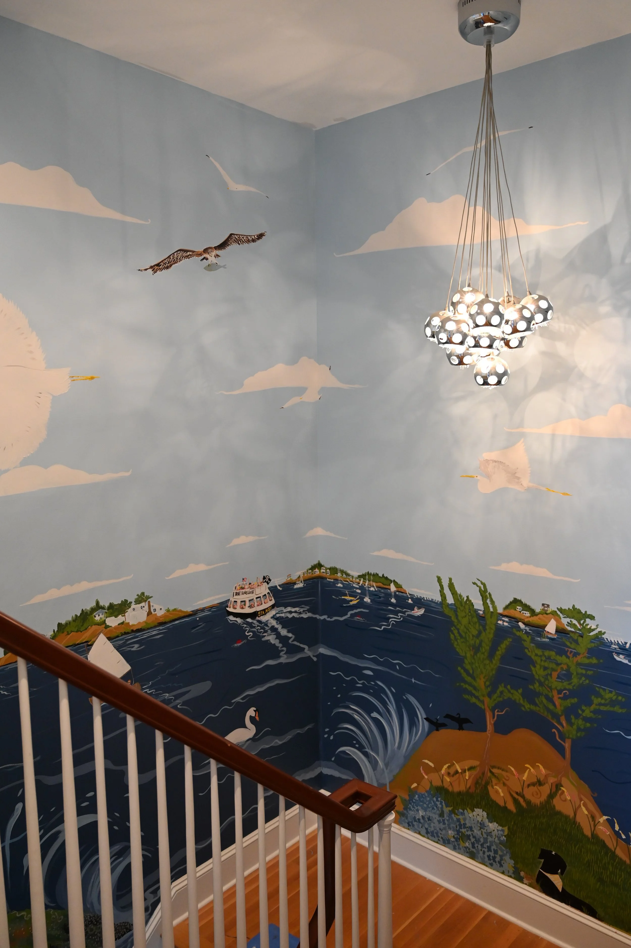 Interior of a staircase with a mural of a water scene, including a boat, a swan, and trees, on the wall. A modern chandelier with multiple black and white spherical lights hangs from the ceiling.