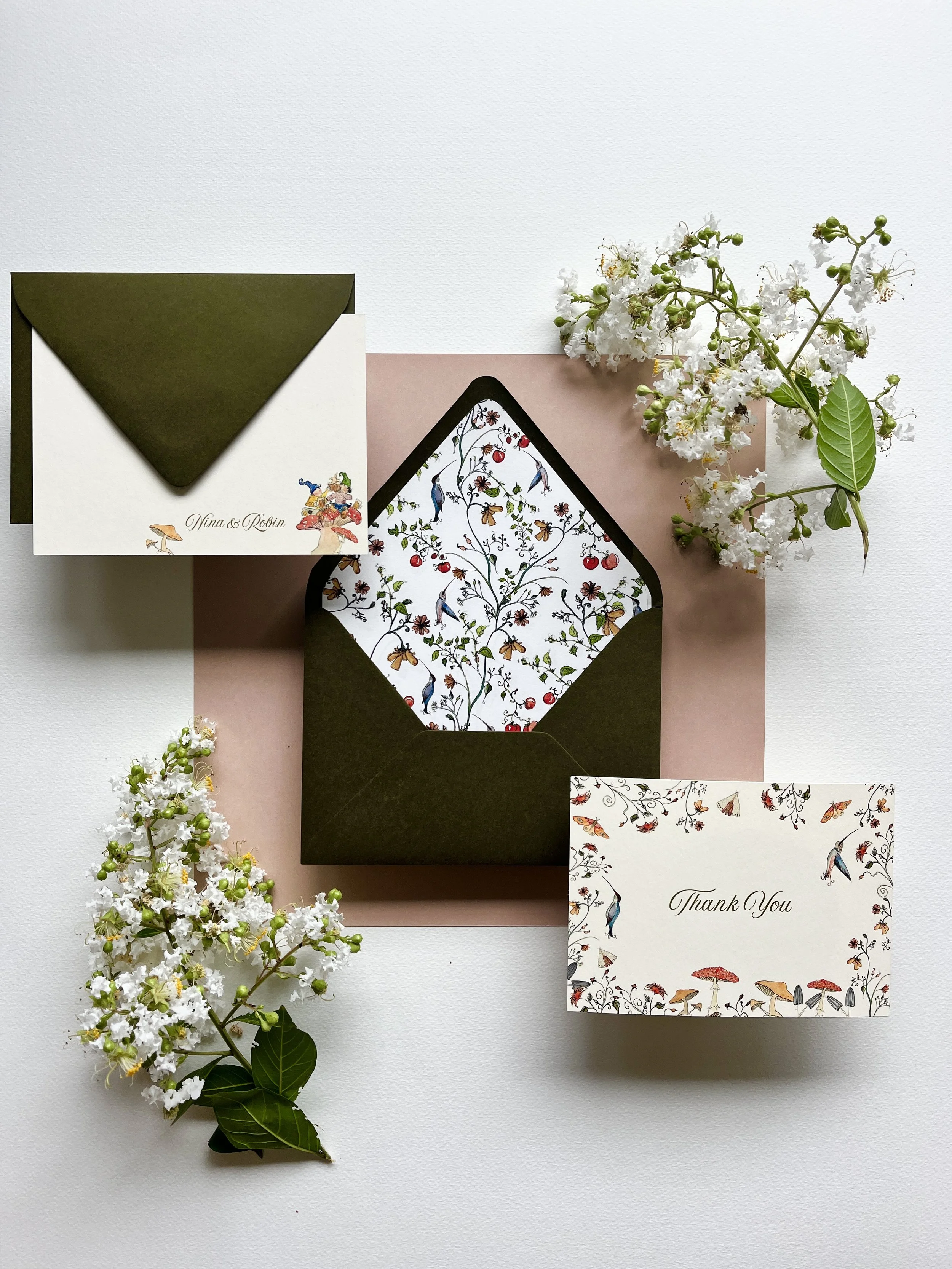 An open thank you card with floral and bird patterns inside, placed in an olive green envelope on pink paper, surrounded by white flowers and a matching card with thank you message.