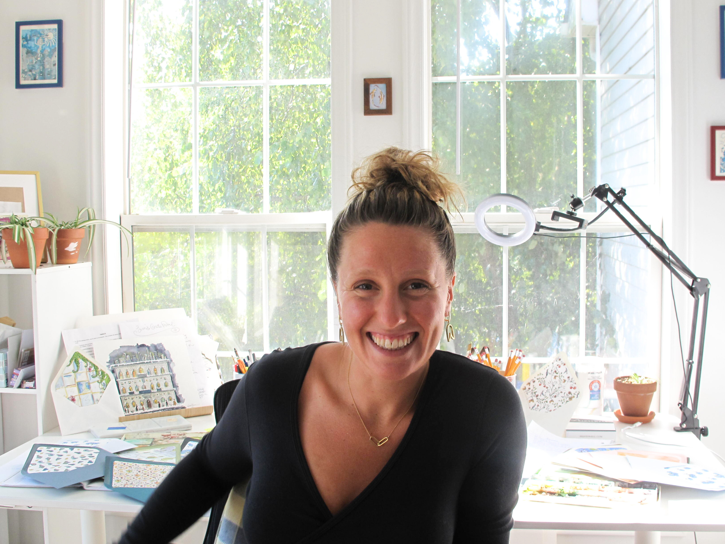 A woman with curly hair tied up in a bun, smiling and sitting at a desk in a bright room with large window. The room has artwork, potted plants, notebooks, and art supplies.