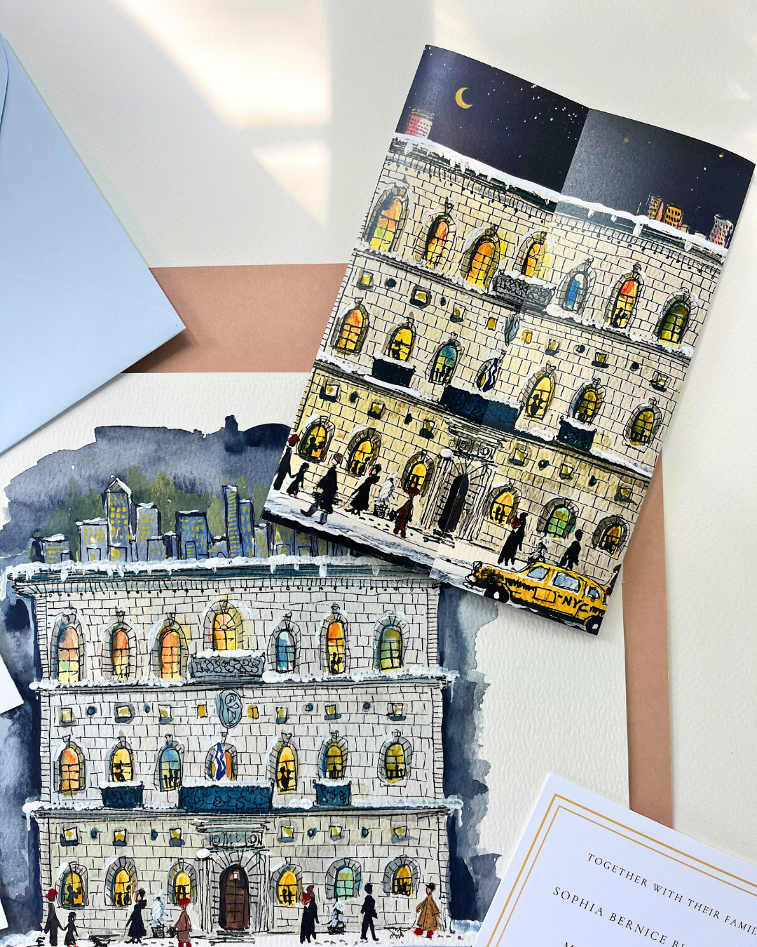 Watercolor paintings of city buildings, depicting a night scene with illuminated windows, people walking on the street, and a yellow taxi in front of a historic building. A plain invitation card is partially visible in the bottom right corner.