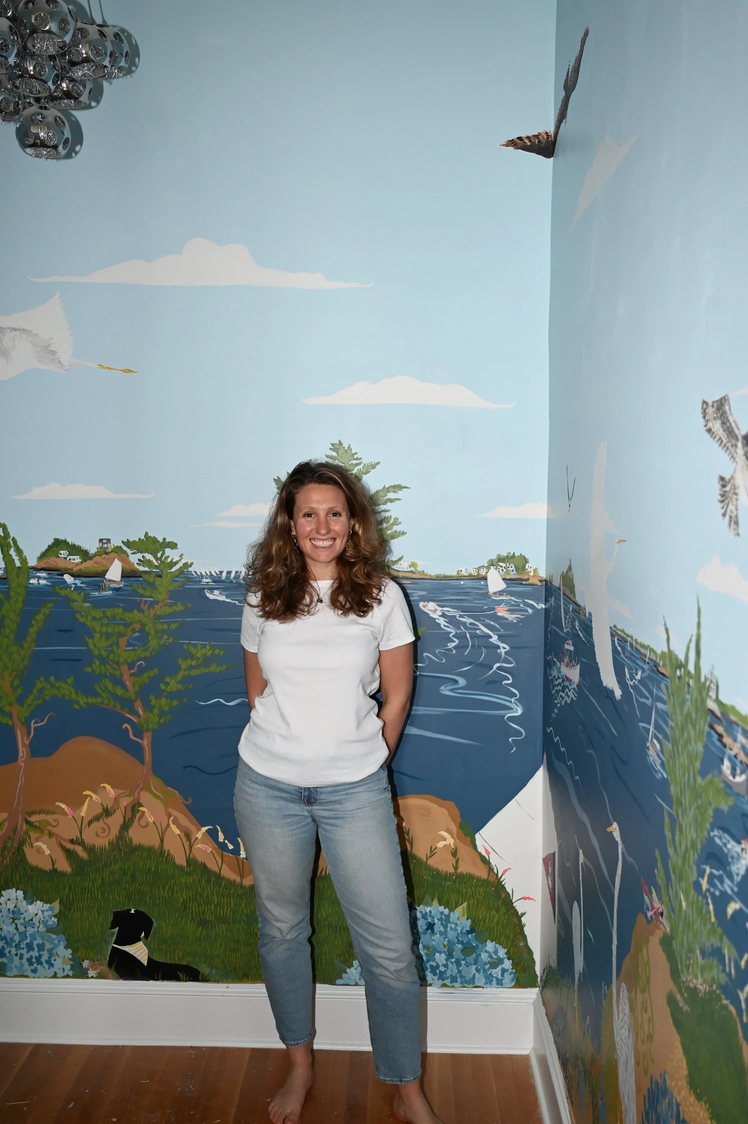A woman with curly brown hair smiling, standing barefoot in front of a painted mural of a coastal scene with water, trees, birds, and sailboats on a light blue wall.