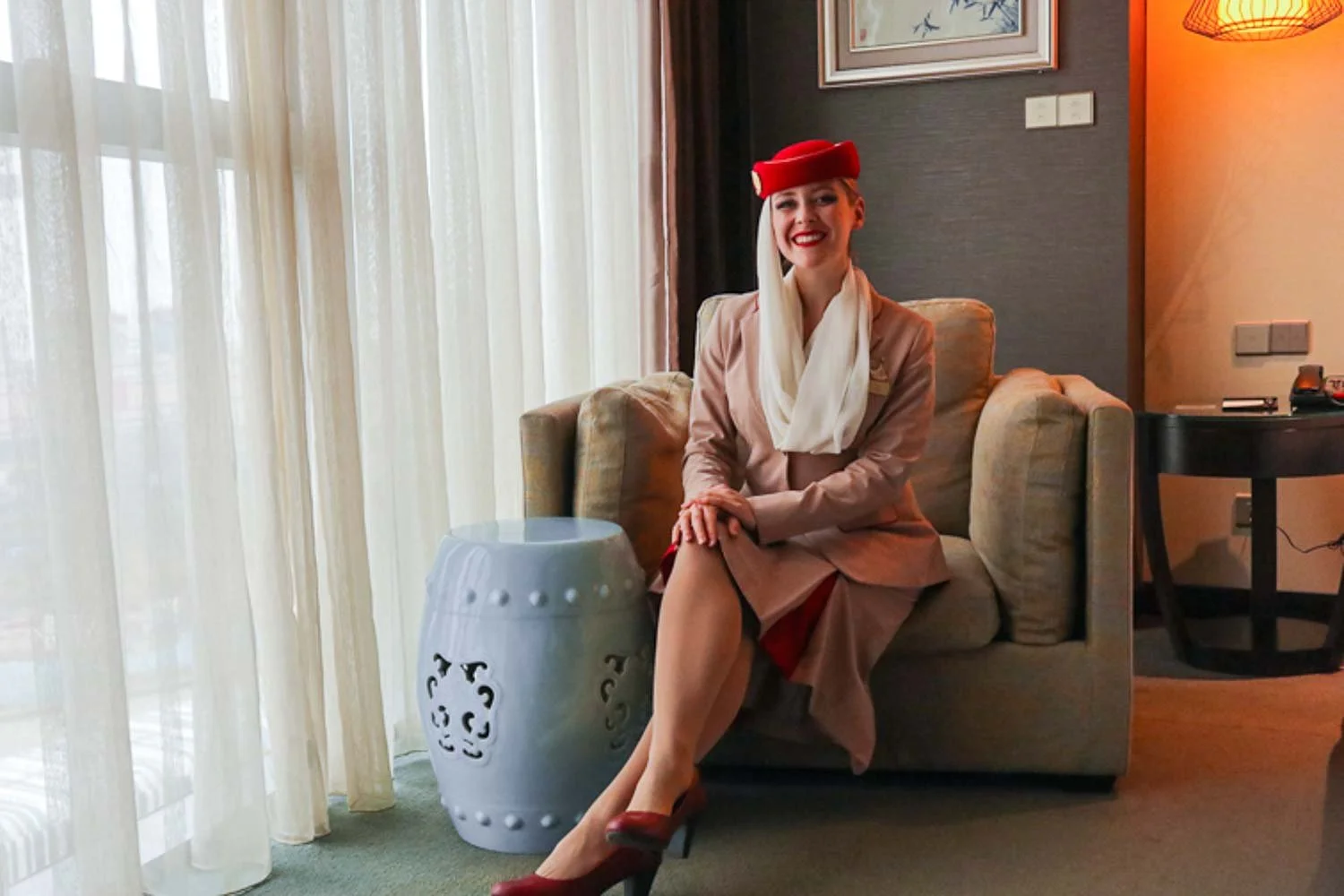 Mrs Timcsi (Csürös Tímea) in Fly Emirates cabin crew uniform joyfully and dynamically leaning forward, while sitting on a stunning armchair in a Shanghai luxury hotel room. From the window on the left the daylight beautifully fills the room.