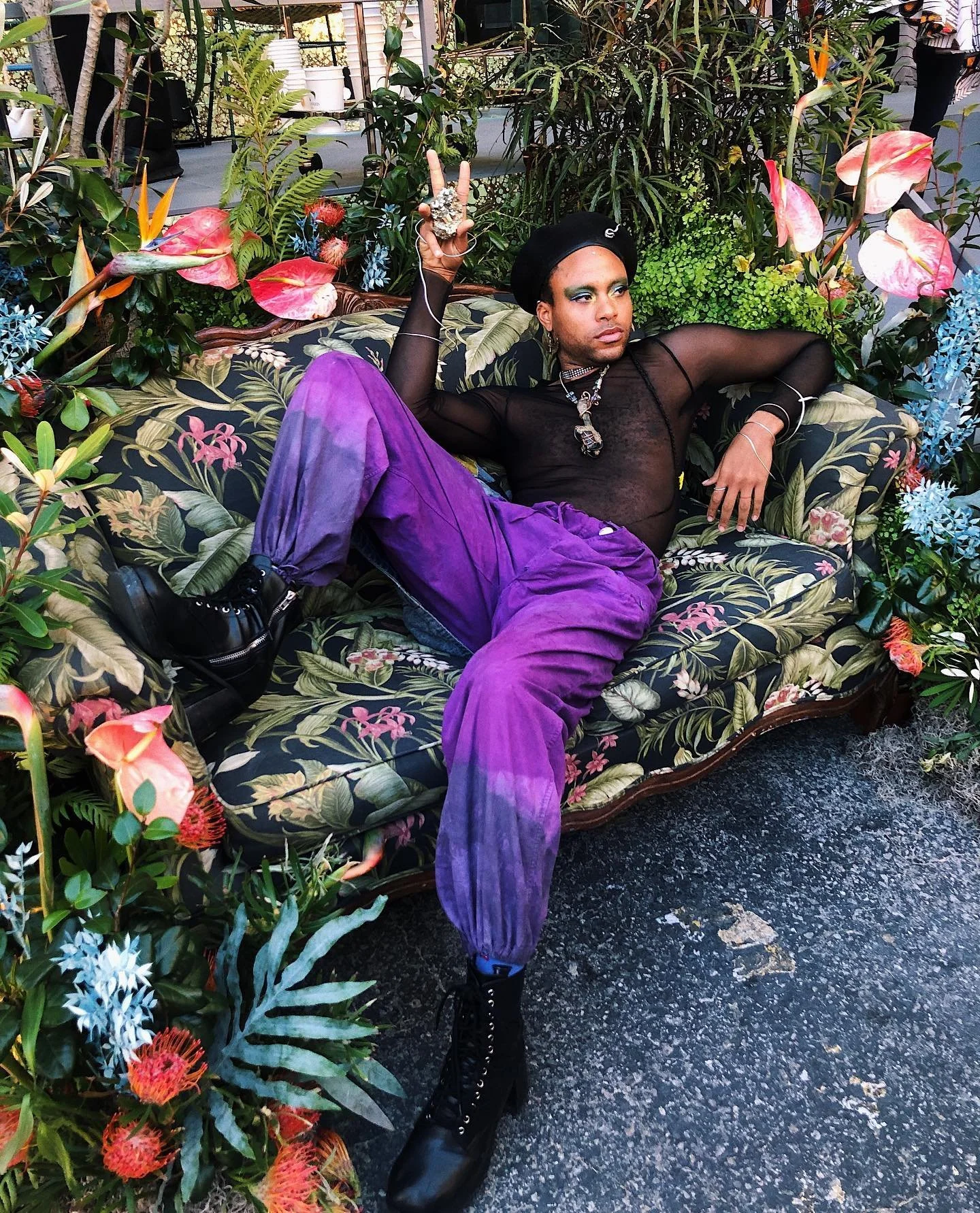 black queer person dressed in a mesh black shits and purple dyed pants holds a piece of gold pyrite surrounded by green plants and flowers