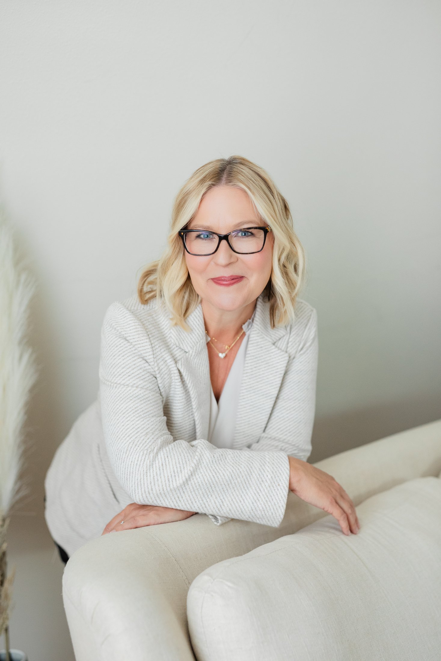 Professional studio portrait of Ann wearing a light blazer while leaning on a light linen sofa, celebrating beauty and confidence after 50.