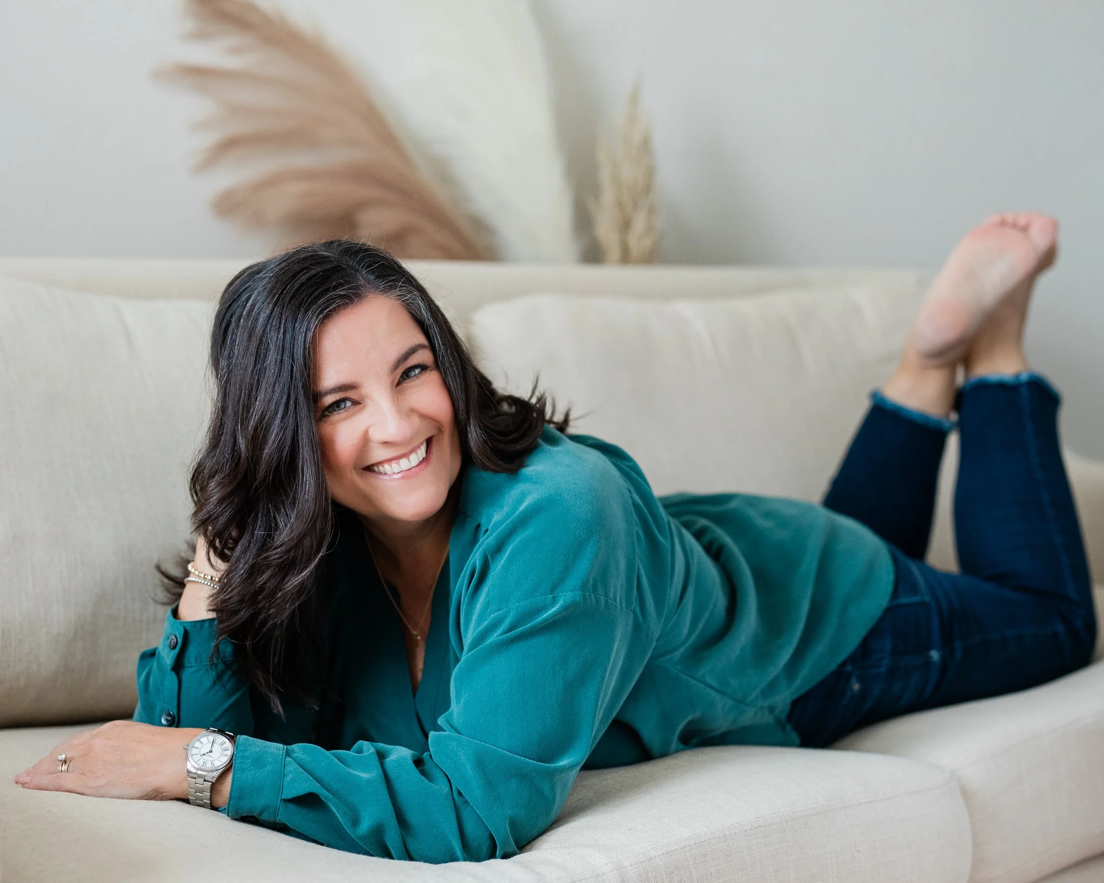 Studio Portrait of a woman over 50 wearing a green blouse and blue jeans, lying on a white sofa. By Amanda Faucett Photography in Houston, TX