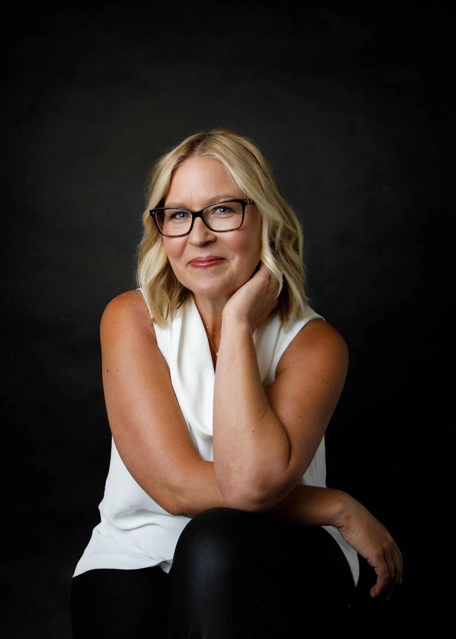 Timeless studio portrait of Ann on a black backdrop wearing a white tank top and black leather pants, celebrating beauty and confidence after 50.