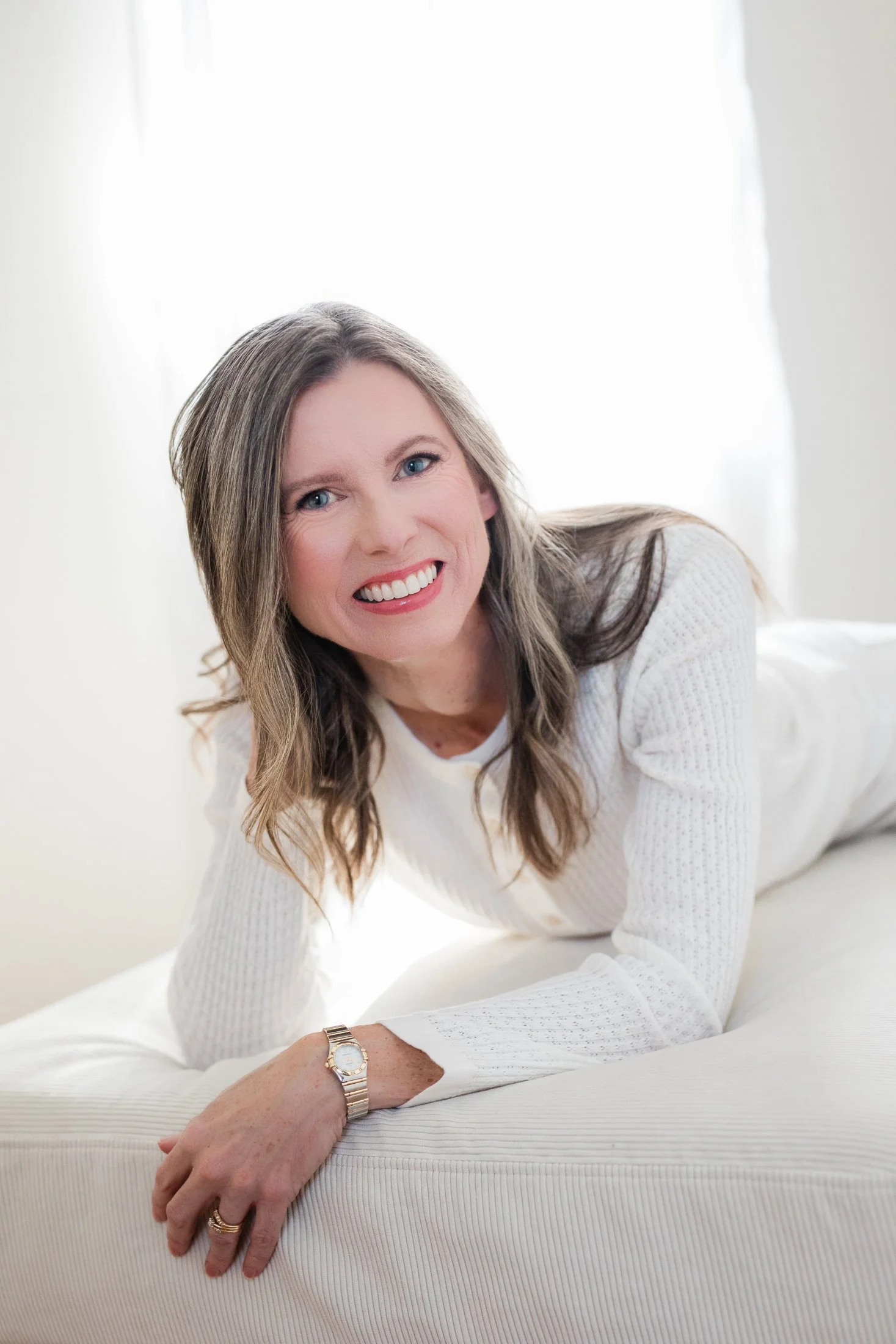 Studio Portrait of a woman over 50 wearing a white top and white jeans, lying on an ottoman by Amanda Faucett Photography in Houston, TX