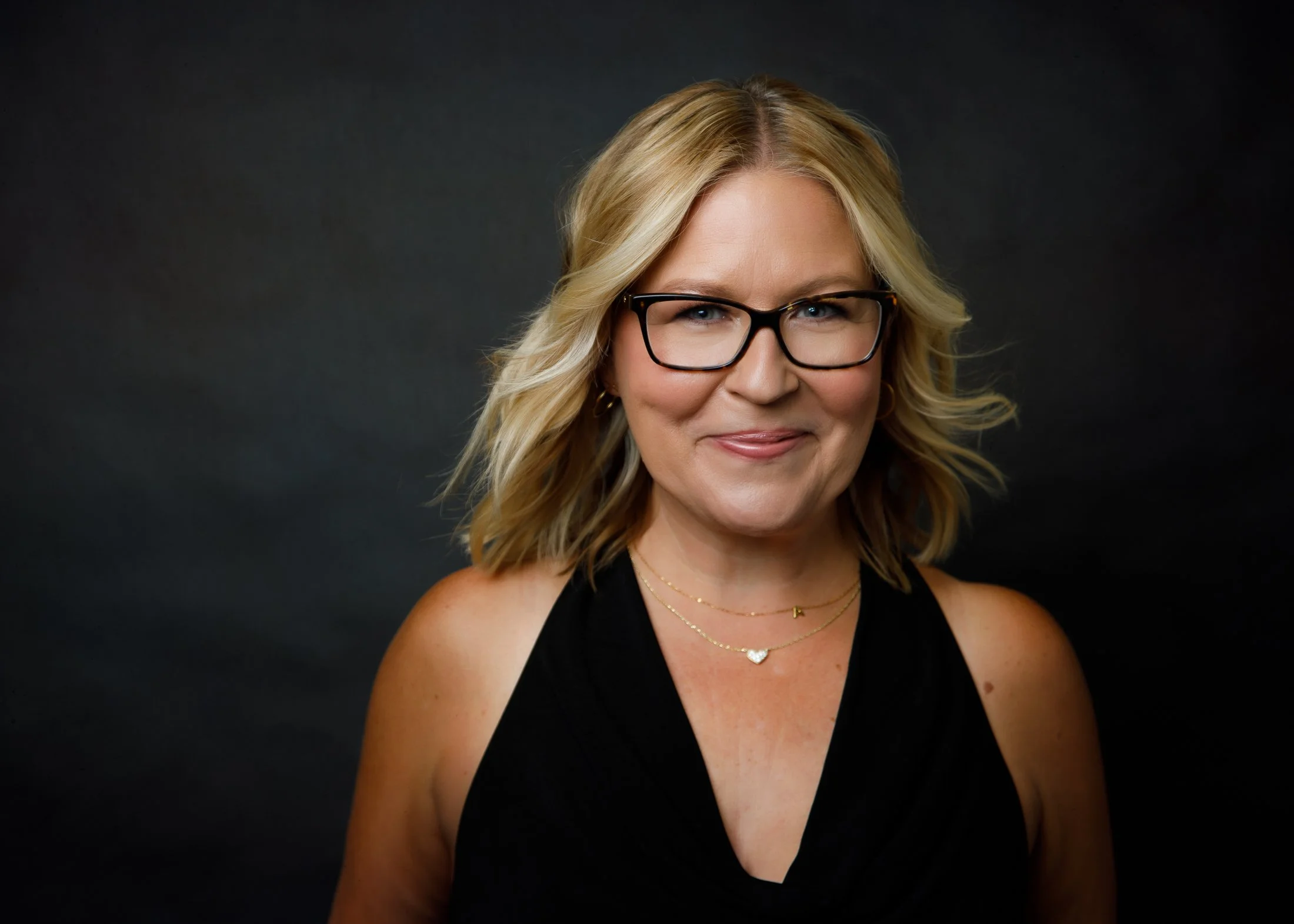 Elegant portrait of Ann on a black background with a black tank top, wearing glasses, from the Wise Women 50 Over 50 Portrait Experience in Houston by Amanda Faucett.