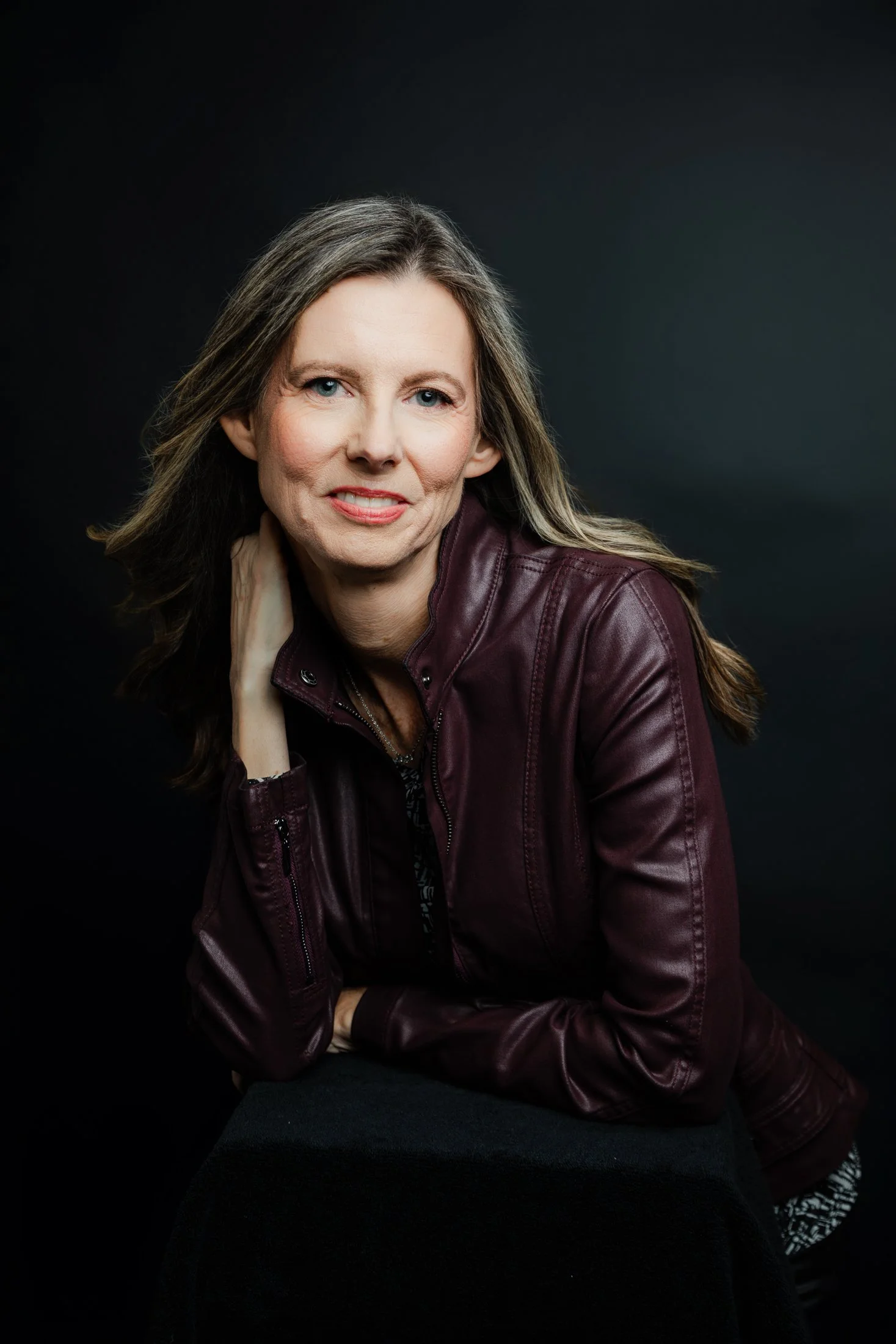 Studio Portrait of a woman over 50 wearing a maroon leather jacket and black jeans, holding the jacket lapels by Amanda Faucett Photography in Houston, TX
