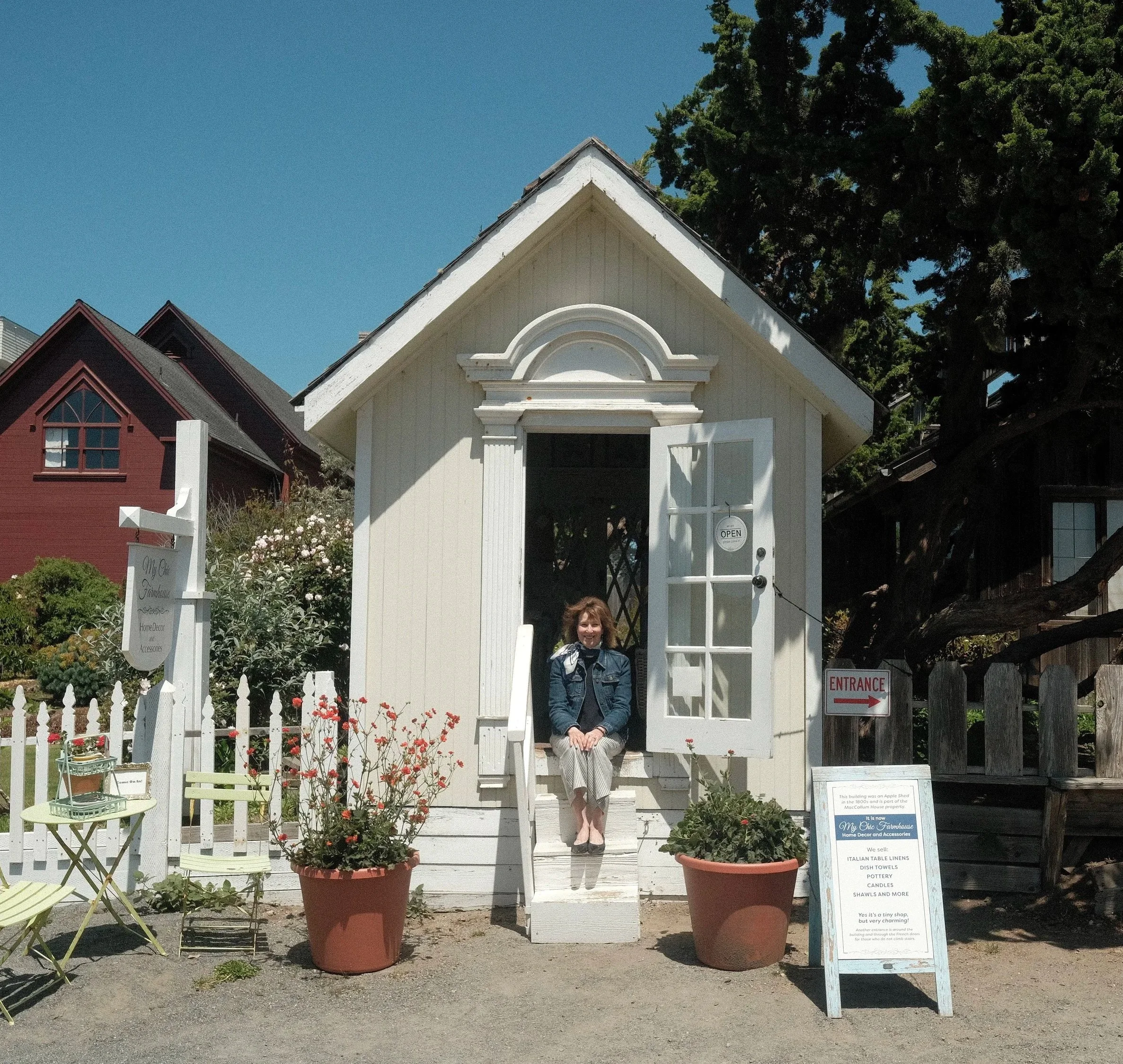 A woman sitting on the porch of a small white building, with a sign promoting home decor and accessories, surrounded by plants and flowers, with a larger red house and trees in the background under a blue sky.